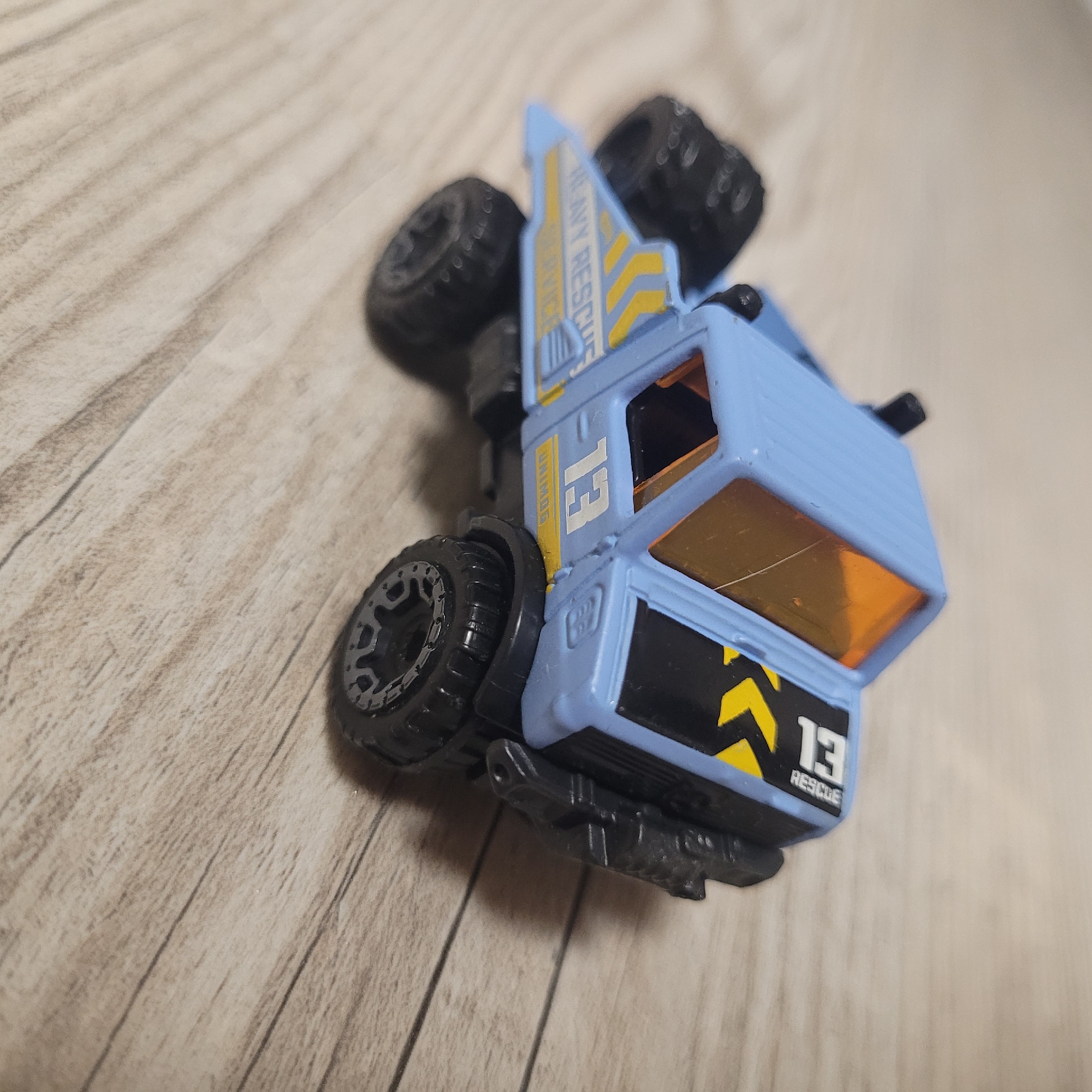 A blue and black toy truck is shown on a wooden surface. The truck is a rescue vehicle with "HEAVY RESCUE" and "SERVICE" written on the side in yellow text. It has the number "13" in white on its side and a black and yellow hazard stripe design. The truck's windows are orange. The wooden surface has a light brown wood grain pattern.