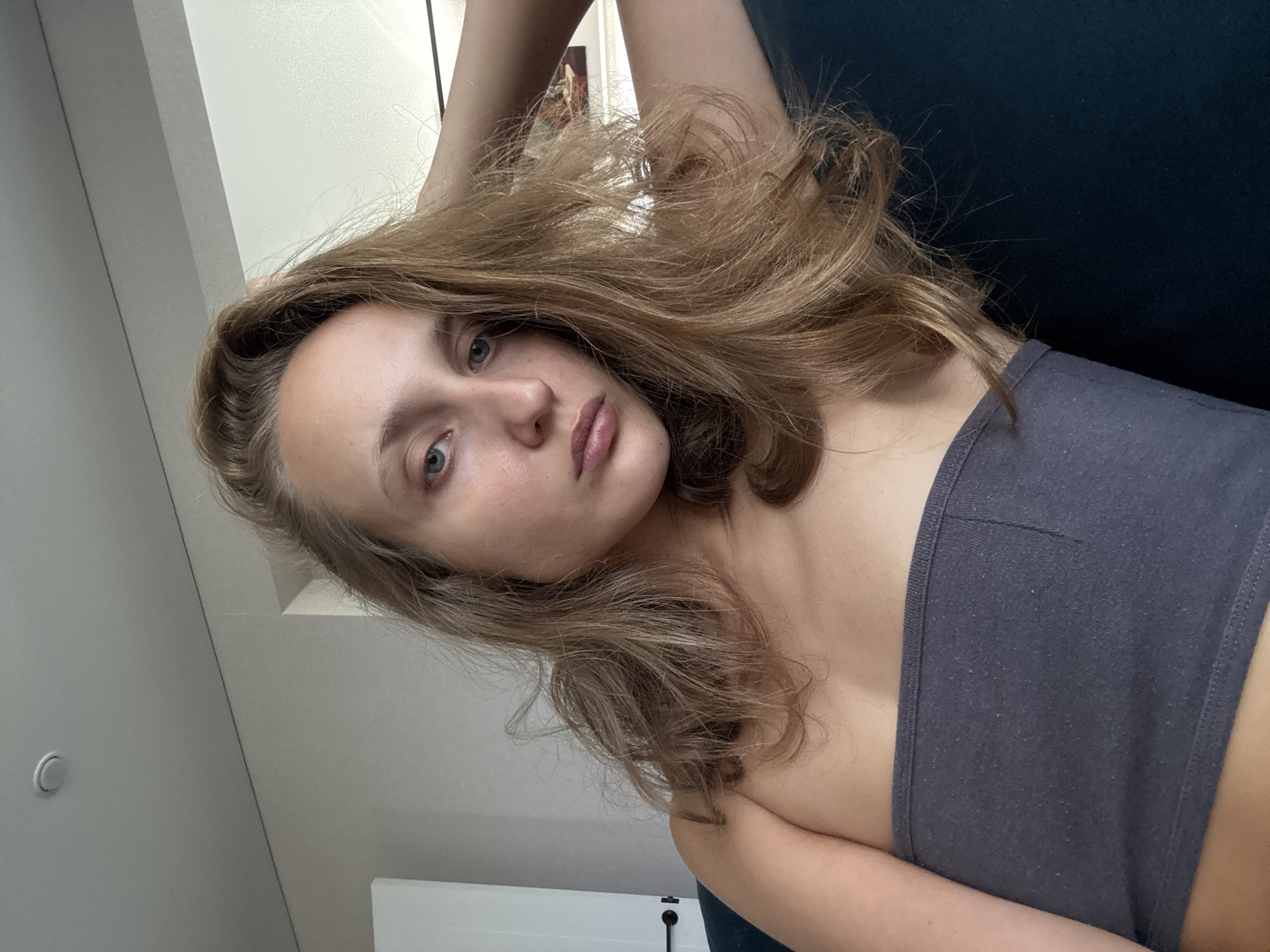 A young woman with long, blonde hair and blue eyes is captured in a close-up, waist-up, indoor selfie. She is wearing a gray strapless top and appears to be reclining on a dark teal couch. Her right arm is bent, with her hand resting on the back of her head, and her hair is draped over her shoulder and arm. The background includes a lightly colored wall, a door with a black handle, and a framed picture on the wall. The lighting suggests it is daytime, possibly near a window. The location is stated as Podshivalovo, Russia.