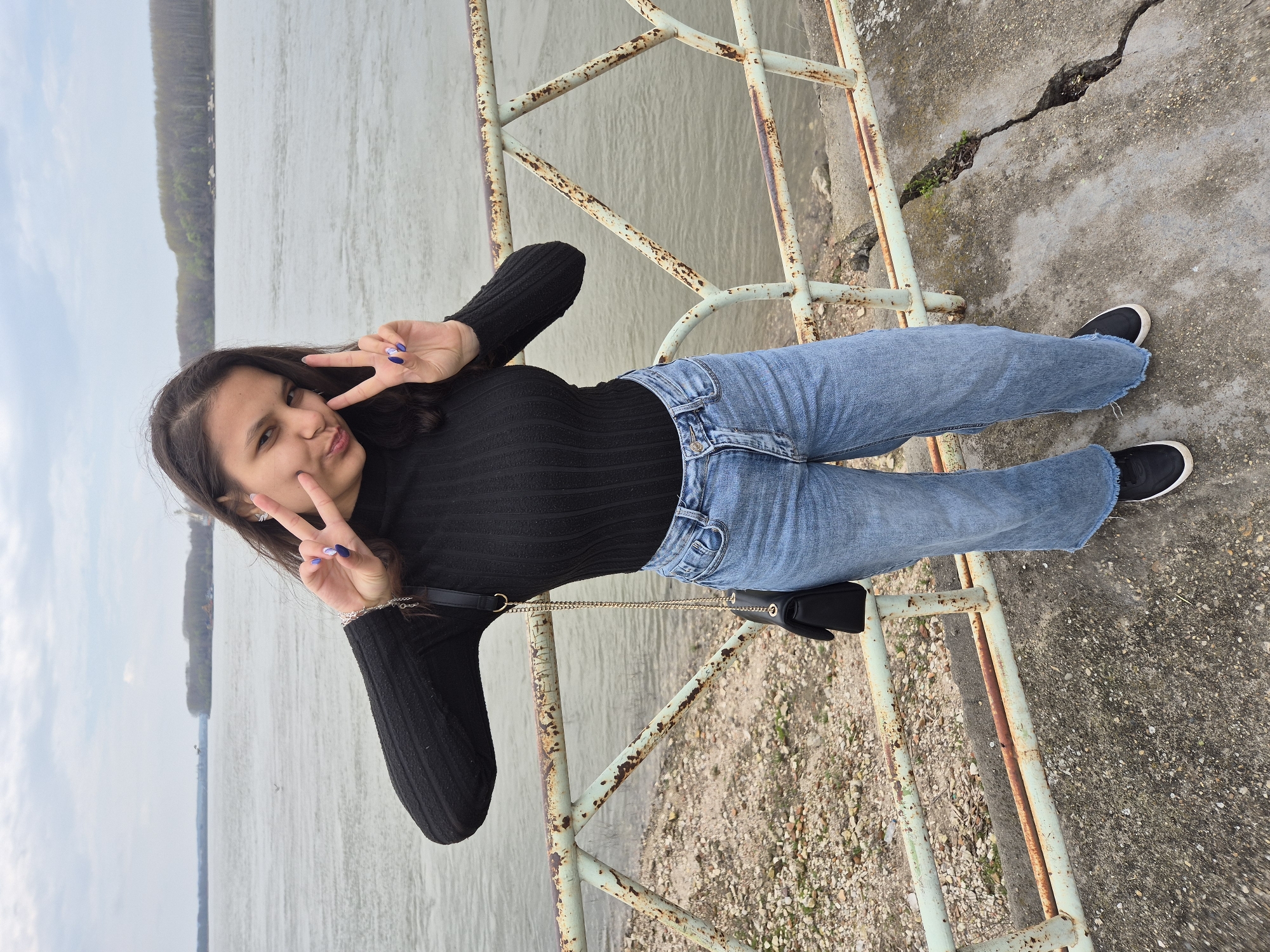 A young woman stands on a concrete platform near a river, likely the Danube, based on the location of Silistra, Bulgaria. She poses, making the "peace" sign with both hands, her nails painted a vibrant blue-purple. She wears a black long-sleeved top, blue jeans, and black shoes. A small black bag with a chain strap hangs on her side. The platform is bordered by a rusted metal railing with a pale green tint. Behind her, the river extends to the horizon, with trees visible on the opposite bank. The concrete platform shows signs of wear and cracking.