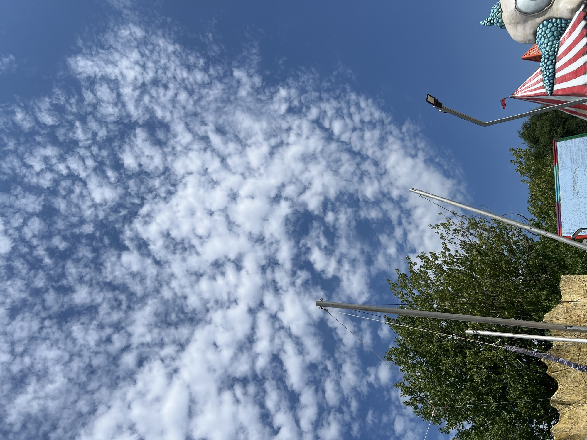 The image shows a sunny day in Golubitskaya, Russia, with a bright blue sky filled with scattered, puffy white clouds. In the foreground, there are parts of amusement park structures, including a large, yellow, wavy-edged object on the left, likely part of a ride. Metal poles and rigging are visible, suggesting some sort of outdoor attraction. To the right, a striped red and white tent peak rises, adorned with a small red flag. Further to the right, a portion of a large, decorative structure with turquoise scaled elements and a prominent eye is visible, suggesting it's part of a themed attraction or mascot. Lush green trees are present in the middle ground, contributing to the outdoor, recreational atmosphere. A street lamp on a pole is also visible in the upper right quadrant. No people are discernible in the image.