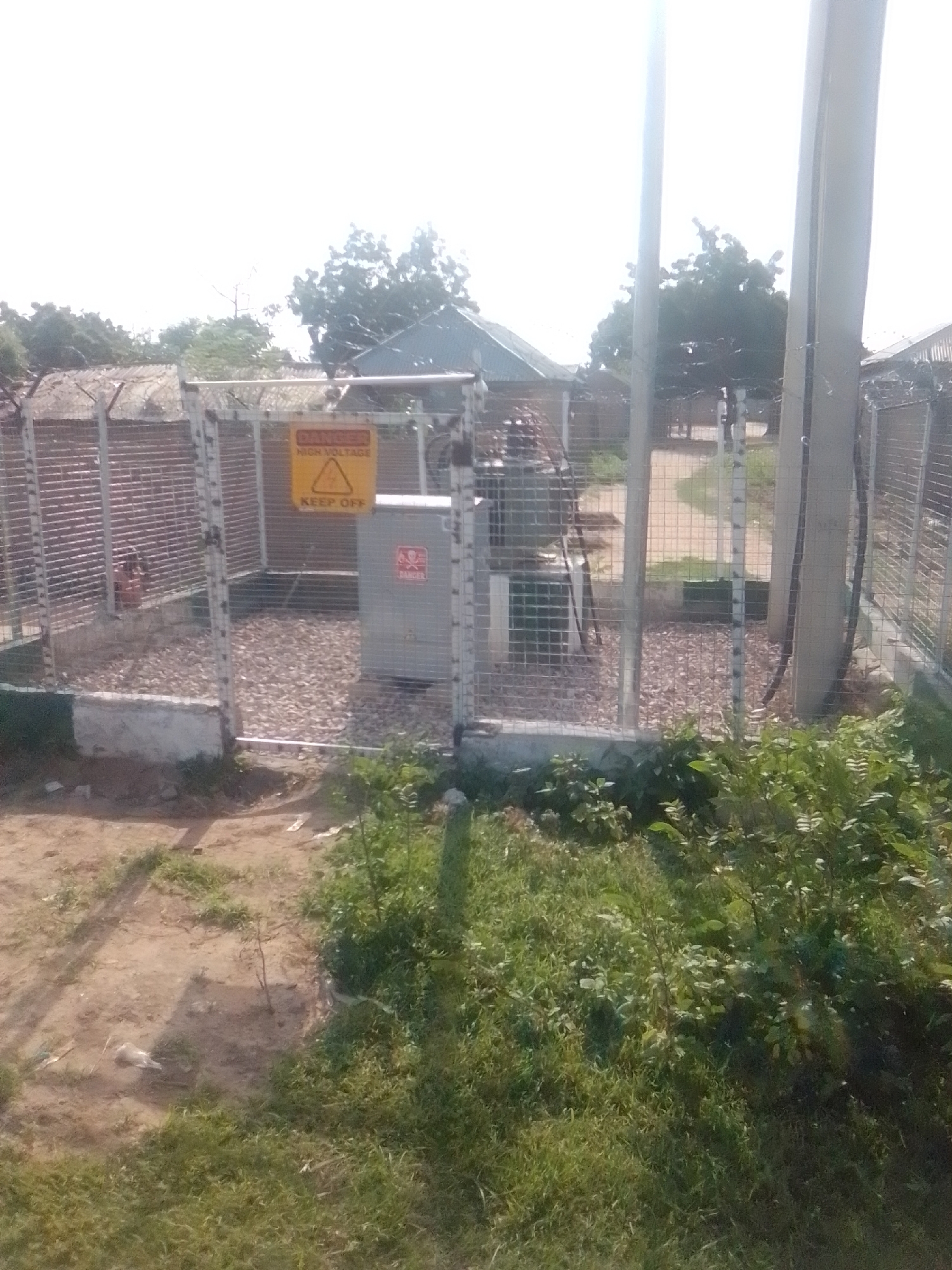 The image shows an outdoor scene in Talasse, Nigeria, depicting an electrical transformer substation enclosed by a chain-link fence. A yellow sign with a lightning bolt symbol and the words "DANGER HIGH VOLTAGE KEEP OFF" is prominently displayed on the fence. A gray transformer box is visible inside the fenced area, resting on a bed of gravel. The substation is surrounded by a concrete base, and beyond the fence, there are trees, buildings with corrugated metal roofs, and more chain-link fencing. The foreground of the image shows patches of grass and dirt. The sky is bright and overcast.