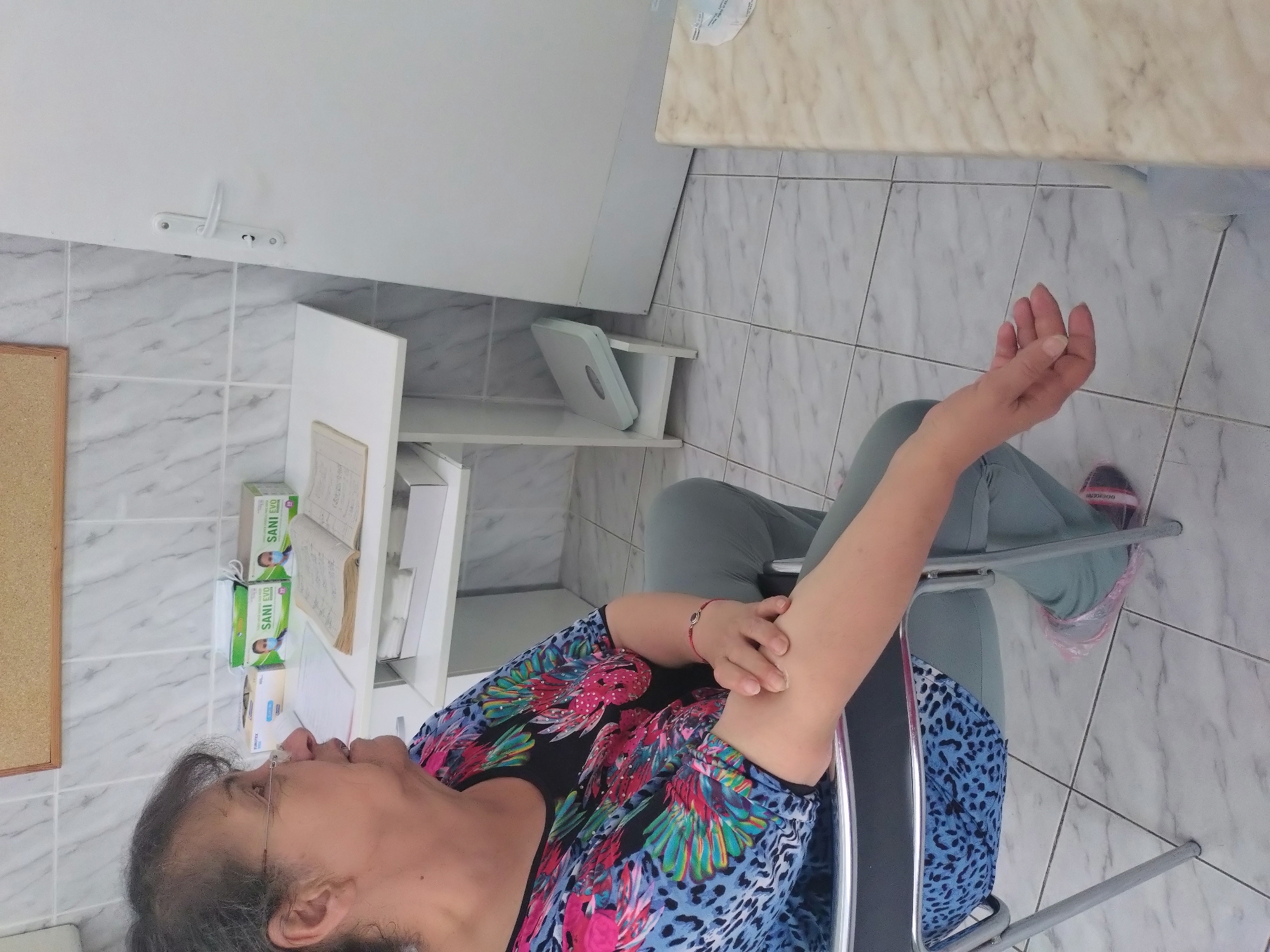 An indoor, eye-level shot depicts an adult woman seated in what appears to be a medical clinic or healthcare facility in Stara Zagora, Bulgaria. The woman, who has dark, graying hair and wears glasses, is dressed in a colorful short-sleeved top with a vibrant pattern of blue, pink, green, and black, and light-colored pants. A red bracelet is visible on her left wrist. She is seated in a metal-framed chair with a blue leopard-print seat and backrest. Her left arm is extended forward, with her right hand gently touching her left forearm, suggesting either discomfort, examination, or preparation for a procedure like a blood test or vaccination.
The background consists of white walls, with the lower half featuring white and gray patterned tiles. To the left, a corkboard is partially visible. Behind the woman, a white shelving unit holds several items, including boxes labeled "SANI EVO," which are clearly face masks given the accompanying icon. Another box, partially visible, reads "CHEMSTEX," likely indicating gloves. An open book or large notebook also rests on the shelf. On a lower shelf of the unit, a white personal scale is positioned. A white wall hook with two prongs is mounted on the wall. The flooring is consistent with the tiled pattern seen on the walls. Her feet, wearing pink-accented shoes, are visible, with what appears to be a clear plastic covering over her right shoe. The neutral expression on her face and the presence of medical supplies reinforce the clinical setting.
