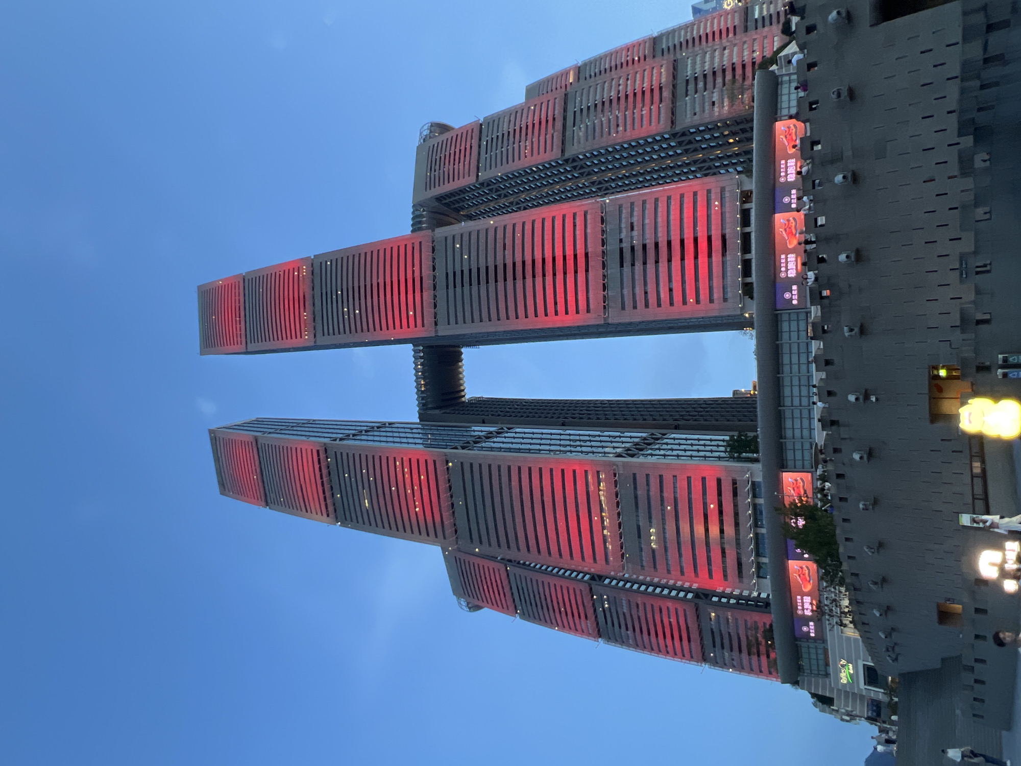 The image shows a low-angle view of a modern skyscraper complex in Yuzhong District, China, at dusk. The complex features two prominent, interconnected towers, each with numerous horizontal windows and vertical facade elements. The buildings are illuminated with bright red lights, creating a striking visual effect against the darkening blue sky. A connecting structure, resembling a horizontal beam or bridge, spans between the two main towers at a high elevation. Below the towers, there is a multi-level structure with a gray, textured facade, possibly a shopping mall or commercial area, featuring large digital screens displaying advertisements. A stone-like wall with decorative elements runs along the foreground, with steps leading up to the elevated area. Several individuals are visible on the steps and along the wall, appearing to be tourists or shoppers. The overall environment suggests an urban setting with notable architectural landmarks.