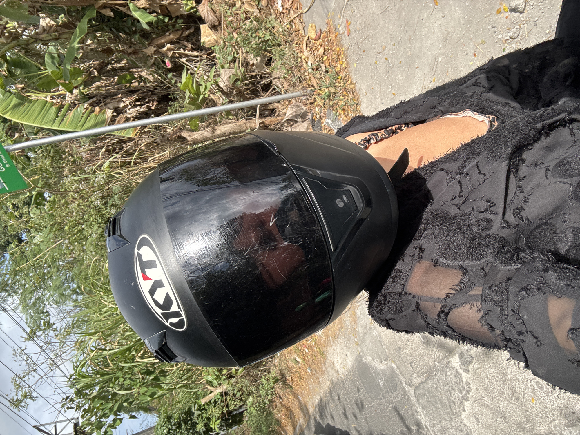 A person wearing a black full-face motorcycle helmet with a reflective visor is positioned in the foreground, obscuring their face. They are also wearing a black sheer, open-weave jacket with a distressed or frayed texture over a patterned top. A necklace with a chain of black and orange links is visible around their neck. The person appears to be seated, possibly on a motorcycle, indicated by the downward angle of the shot and the texture of the pavement beneath them.
The background depicts an outdoor scene with lush green vegetation, including tall grasses and banana-like plants, as well as trees with dry leaves on the ground. A gray pole with a green sign attached to it is visible in the right background, suggesting a roadside environment. The pavement in the foreground is made of interlocking rectangular stones or bricks. The lighting suggests it is daytime, with sunlight casting shadows. The overall environment appears to be a rural or suburban area with abundant plant life.