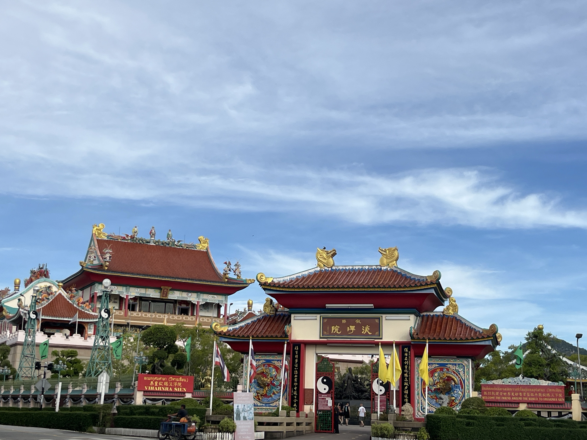 The image shows the entrance to Viharnra Sien, a Chinese temple complex located in มาบฟักทอง, Thailand. The entrance features a traditional Chinese gate with a red tile roof adorned with golden dragon sculptures. The gate is flanked by Thai and yellow flags, and the entrance is decorated with yin-yang symbols. Beyond the gate, there are sculptures. In the background, a multi-tiered Chinese building with a red roof can be seen. The sky is blue with scattered clouds. In the foreground, there are green hedges, a street, and a sign with Thai and English text welcoming visitors to Viharnra Sien. There is a three wheeled vehicle on the left.