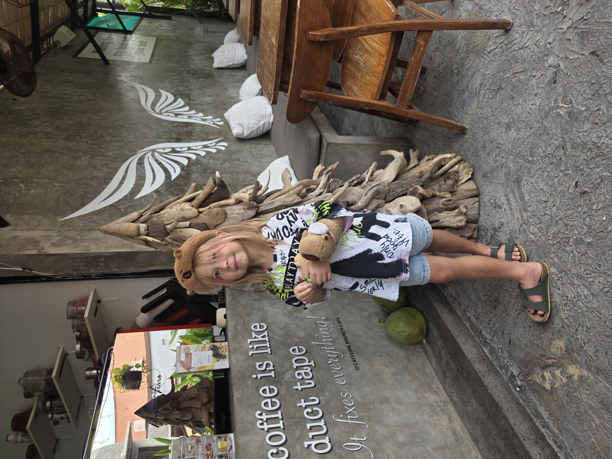 A young girl with blonde hair, wearing a brown bear-shaped hat, a graphic t-shirt, and denim shorts, stands in front of a concrete wall at a cafe in North Kuta, Indonesia. She holds a stuffed toy. To her left, a sign on the wall reads "coffee is like duct tape. It fixes everything!". Two green coconuts are placed on the ground to her left. Behind her, a large, abstract sculpture made of driftwood stands upright. The cafe setting includes wooden tables and chairs, white cushions on a bench, and shelves with jars and decorative items. The overall environment suggests an outdoor or semi-outdoor seating area.