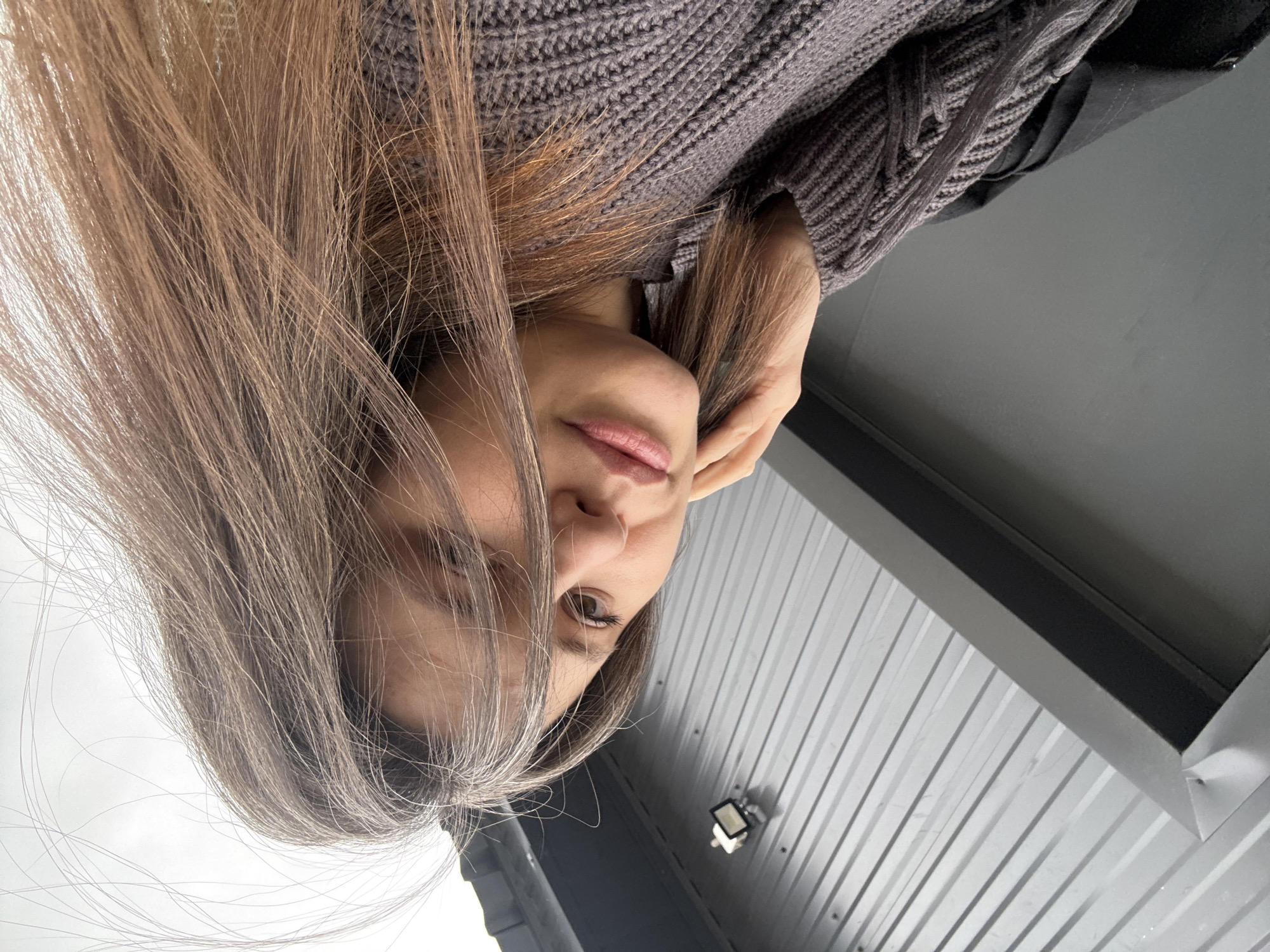 A woman with long brown hair is positioned indoors, in front of a corrugated metal ceiling. She is wearing a grey knit sweater. The location is Chaikovsky, Russia. There is a light fixture on the ceiling.