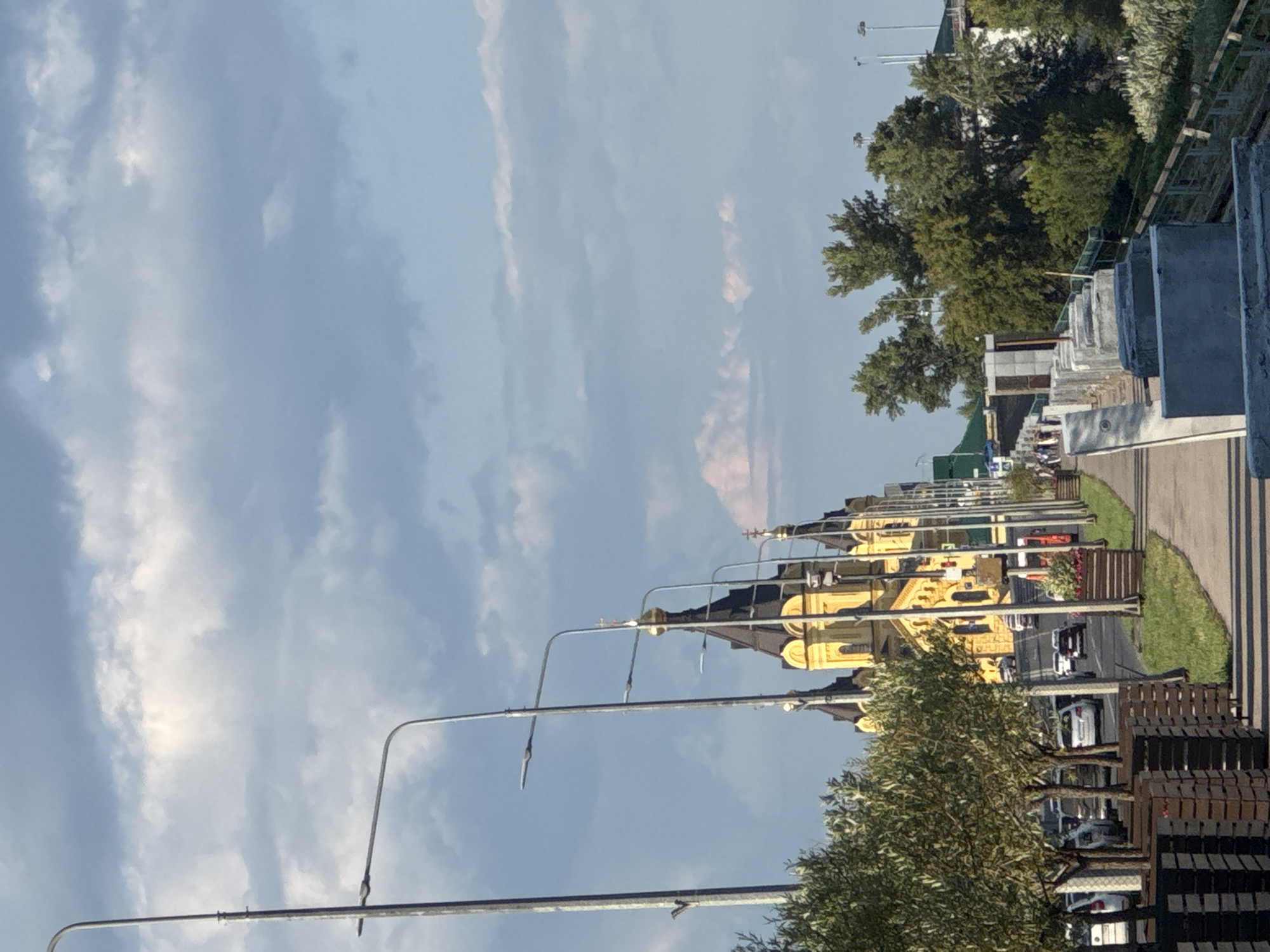 The image shows a view from a sidewalk looking towards a street and a prominent yellow church with black domes, identified as the Alexander Nevsky Cathedral in Nizhny Novgorod, Russia. The sidewalk is lined with trees in wooden planters on the left and a low concrete wall with barriers on the right. Cars are visible on the street, with traffic moving. In the background, there are more trees and some buildings. The sky is partly cloudy, with a mix of blue sky and white clouds. A lamppost is also visible on the left side of the image.