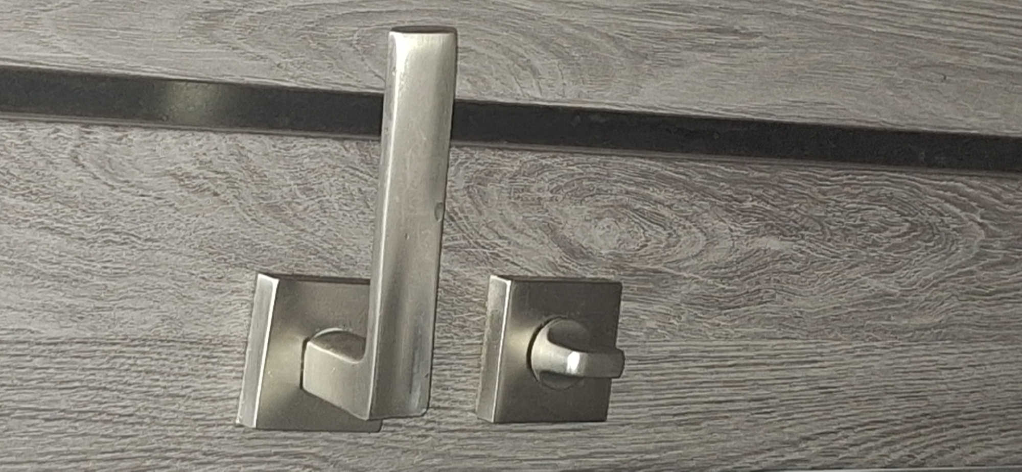 The image shows a close-up view of a brushed metal door handle and lock mechanism on a gray wood-textured door. The door handle is horizontal and rectangular in shape, attached to a square base. Below the handle is a square lock escutcheon with a circular knob. The wood grain on the door is prominent, with a vertical orientation. The scene appears to be an interior door, possibly in a residential or commercial building. There are no people or actions depicted in the image. The location in Bila Tserkva, Ukraine, is not discernible from the visual information provided in the image.