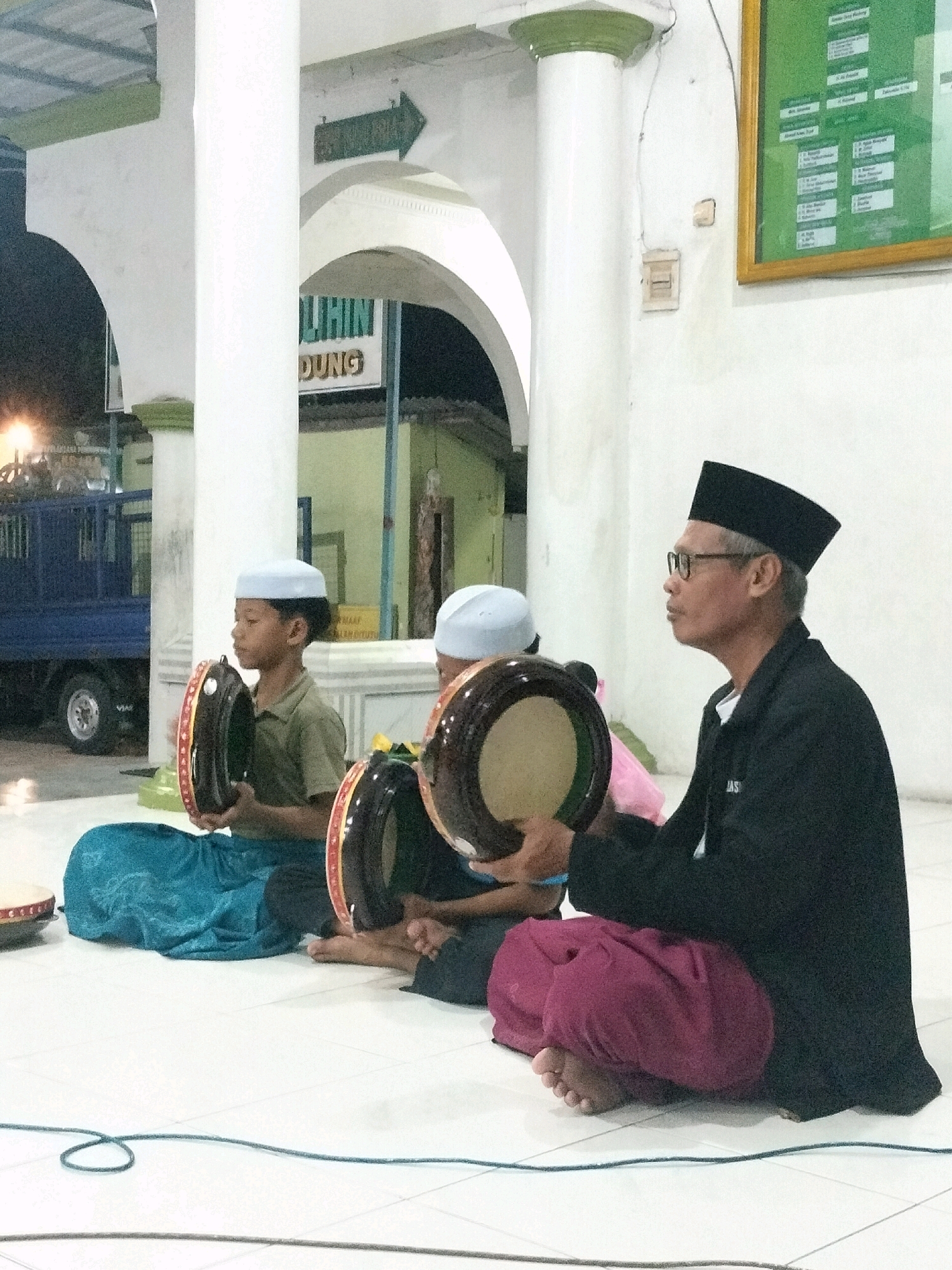 A group of three people, two young boys and an adult male, are sitting on a tiled floor, appearing to be playing musical instruments. The adult male, wearing a black peci hat and glasses, is on the right, holding a tambourine-like drum. The two boys, dressed in light blue peci hats and casual shirts, are positioned to his left. One boy is wearing a turquoise sarong, while the other's attire is partially obscured. Both boys are holding similar drums. The setting appears to be an interior space with white pillars, arched doorways, and a green sign with text that includes "LIHIN DUNG" and an arrow, suggesting it's in Wedung, Indonesia. A blue truck is visible through an archway in the background. The overall atmosphere suggests a cultural or religious gathering, possibly a musical performance.
