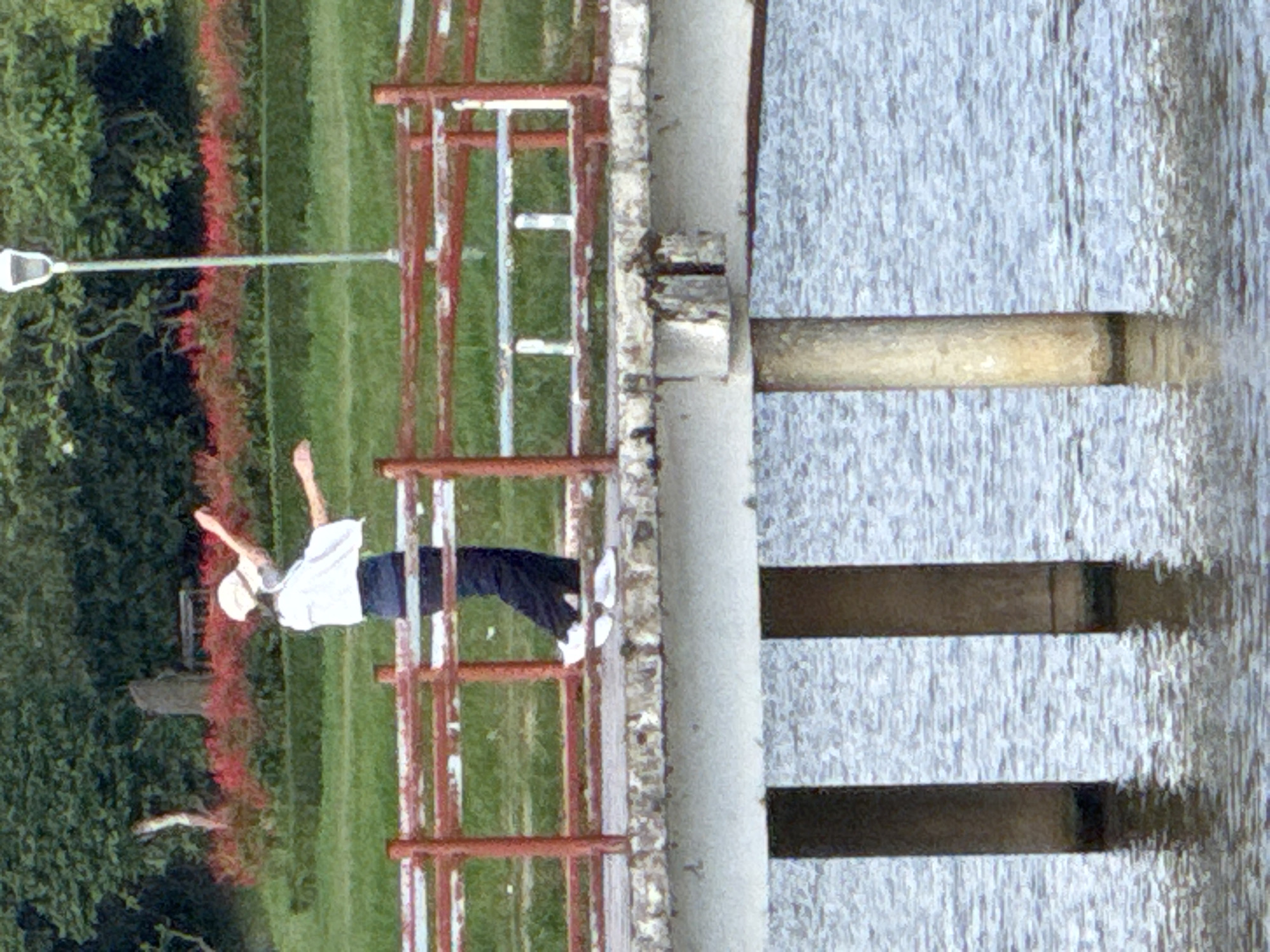 The image captures a scene on a bridge in Phường Xuân Hương - Đà Lạt, Vietnam, overlooking a body of water. A person stands on the concrete bridge deck, slightly left of the image's center, facing towards the right. They are dressed in a white wide-brimmed hat, a loose-fitting white long-sleeved shirt, dark blue trousers, and white athletic shoes. Both arms are raised and extended outwards, with the hands open; a small white object is perceptible near the left hand. The bridge features a two-tier railing system, primarily painted reddish-brown with white horizontal bars on the lower tier. Concrete pillars support the bridge deck, with their bases submerged in the rippled, blue-grey water below. A slender white street lamp with a conical top is positioned on the bridge to the right of the person. The background shows a green grassy bank, followed by a dense hedge of reddish-pink flowering bushes, and further back, a thick canopy of green trees. A light-colored tree trunk and a darker, squat structure are partially visible in the distant foliage.