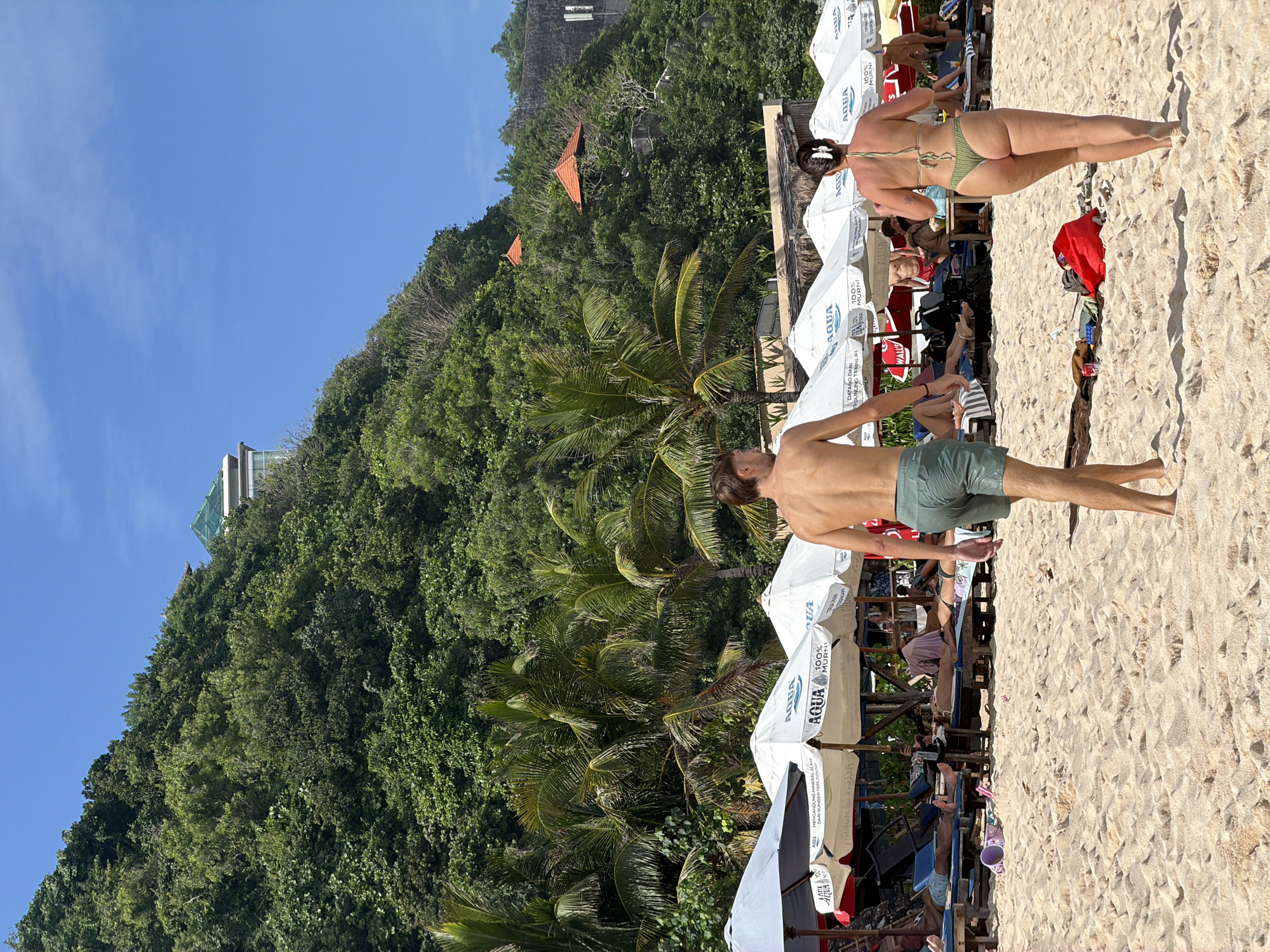 The image captures a vibrant scene at Melasti Beach in Kuta Selatan, Indonesia. A man and a woman are strolling barefoot on the sandy beach. The man is shirtless, wearing green shorts, while the woman is in a green bikini. They are walking near rows of white sun umbrellas and sun loungers where other beachgoers are relaxing. Behind the beach area, a lush green hill rises, dotted with palm trees and other vegetation. In the distance, there are buildings, including a structure with a green roof. The sky above is clear and blue.