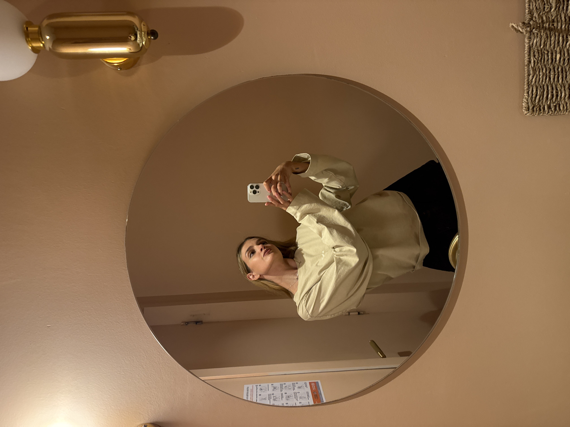 A woman is reflected in a round mirror. She is holding a white smartphone and appears to be taking a photograph of herself in the mirror. She is wearing a light beige, oversized sweater and a black skirt or pants. The room has peach-colored walls. The mirror is mounted on the wall, and the reflection shows a bathroom interior including a door and instructional diagrams posted below the mirror. A gold-colored sconce is visible on the wall to the left of the mirror. A woven basket or piece of art hangs on the wall to the right of the mirror. The location is Tbilisi, Georgia.