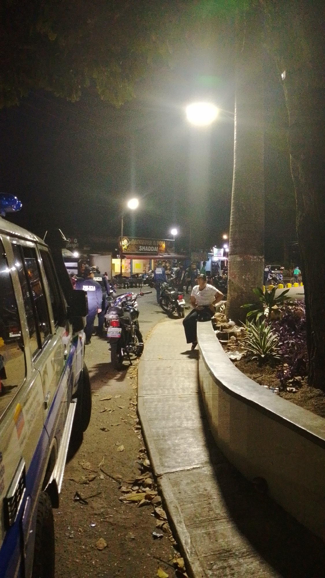 The image depicts an outdoor scene at night in Tinaquillo, Venezuela. Streetlights illuminate the area.
To the left is a light-colored police vehicle, facing away from the viewer. A uniformed officer stands near the vehicle. In the middle ground, several motorcycles are parked along the curb. A man in a striped shirt and dark pants is seated on a concrete retaining wall.
In the background, a restaurant called "Shaddai" is visible. People are seated at tables outside the restaurant. Other people, possibly police officers, are visible in the background. The ground is covered with leaves and debris. Trees are visible along the right side of the image.