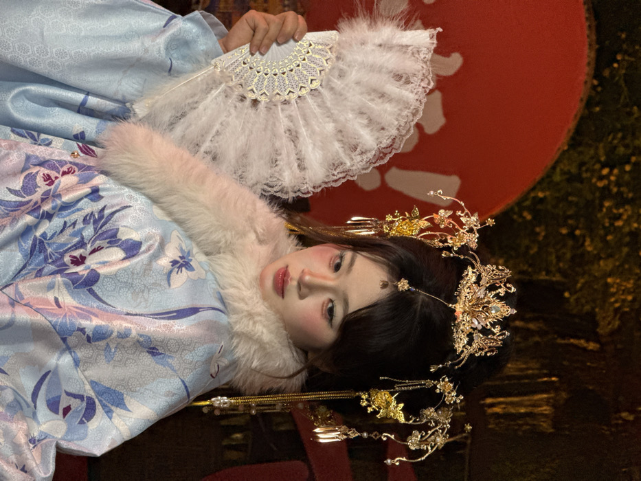 A close-up, chest-up portrait depicts an individual in traditional Chinese attire, positioned in Yuzhong District, China. The subject, appearing female, has dark hair styled into an ornate updo. A multi-tiered golden headdress adorns the hair, featuring intricate filigree, iridescent gemstones, and dangling elements including chains, beads, and butterfly motifs, with a central drop on the forehead. The individual's face exhibits prominent pink blush on the cheeks, light green eyeshadow, and light-colored lips, with the gaze directed downward and to the left. She wears a light blue traditional garment, possibly silk or satin, patterned with large floral designs in shades of blue, purple, and pink, over a subtle lighter blue geometric background pattern. A thick, off-white fur collar encircles her neck and shoulders. Her right hand holds a white feather fan, partially open, displaying a decorative, metallic or iridescent, lace-like frame. The background features a large, bright red, circular or semi-circular object, likely a decorative screen or lantern, directly behind the subject. Beyond this, the environment is dark and indistinct, showing sparse illumination on what appears to be foliage or tree branches, indicative of a nighttime setting.