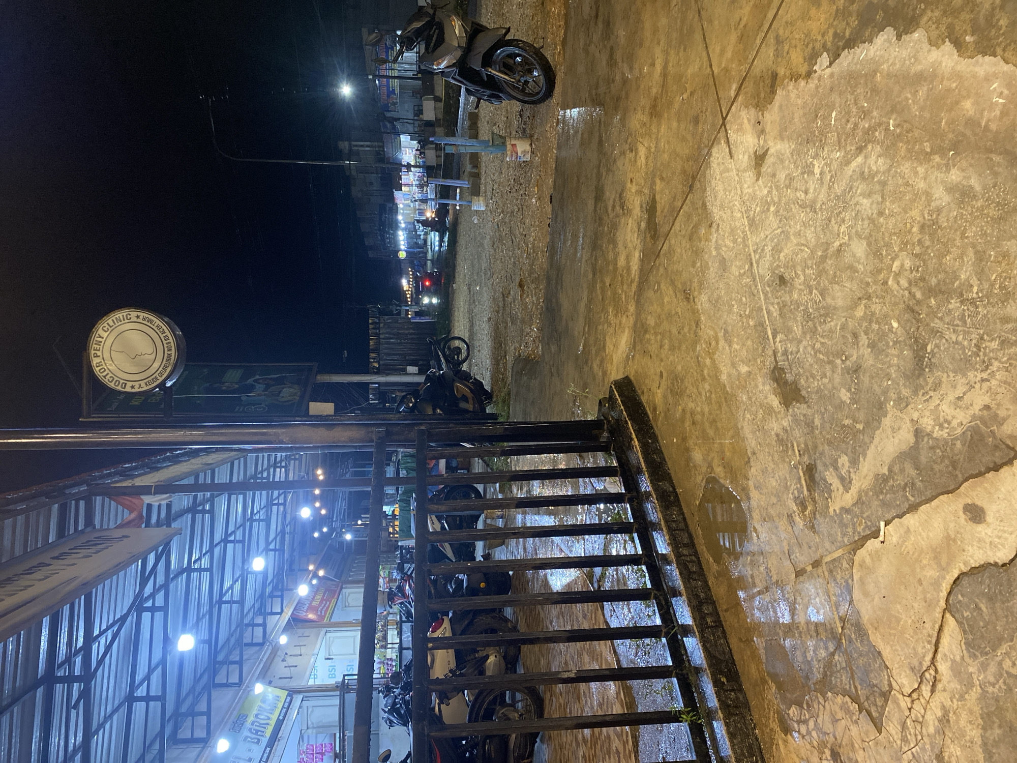 This night-time image depicts a wet street or pathway in an urban or semi-urban setting, likely after rainfall. The perspective is from a slightly elevated position, looking down towards a street lined with shops or stalls.
In the immediate foreground, a dark metal gate or railing extends diagonally across the lower left side of the frame. The concrete ground is visibly wet, showing numerous puddles that reflect the overhead lights. The ground is textured with cracks and various patterns, including what appears to be footprints and debris.
Beyond the gate, several motorcycles, primarily scooters, are parked. A prominent dark grey scooter is positioned on the wet ground in the mid-ground. The area under a large, possibly corrugated metal, awning or roof structure is brightly illuminated by numerous artificial lights. This area appears to house small businesses or market stalls.
Several signs and banners are visible:
- A circular sign, prominently displayed, reads "DOCTOR PENY CLINIC" and below it, "JL. KEUDE GEUDONG ACEH TIMUR", providing a clear location cue for Aceh Timur, Indonesia.
- Other banners are partially visible, with text like "PHARMACY" and "AL-BAROKAH".
- Some signs bear the acronym "BSI", likely referring to Bank Syariah Indonesia.
The sky is dark, indicating it is nighttime. A bright streetlight is visible in the upper right, casting light into the dark sky. Faint lights from other establishments or vehicles can be seen in the distance along the street. No specific individuals are clearly discernible or engaged in any particular activity, suggesting a quiet moment. The wet ground and strong artificial lighting are notable features of the scene.