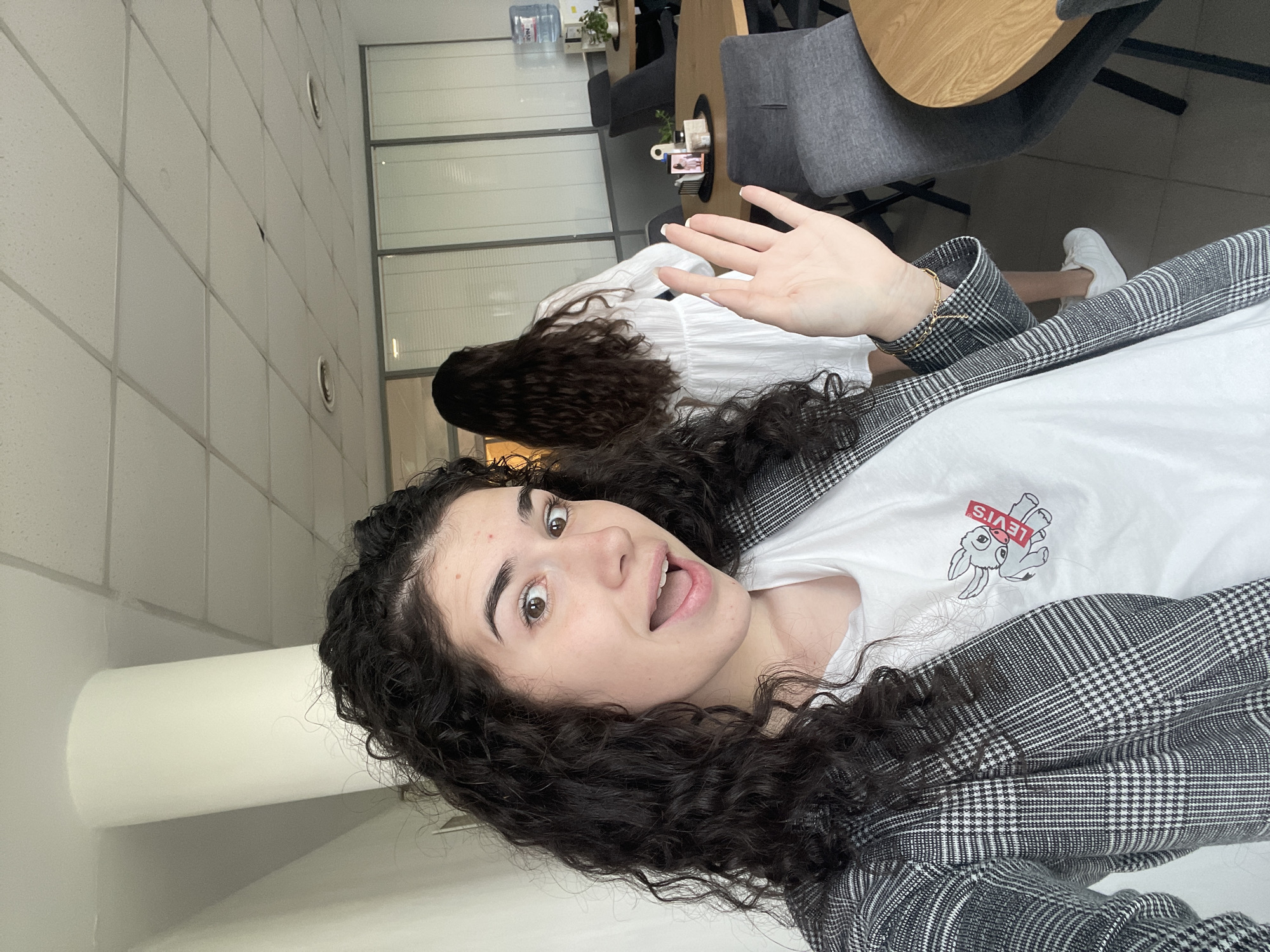 A young woman with curly dark hair is taking a selfie. She is wearing a white t-shirt with a graphic of a rabbit and a "Levi's" logo on it, and a black and white checkered blazer. She is smiling and waving her hand. Another woman with long dark hair, wearing a white dress and white sneakers, is standing in the background. The setting appears to be an office or workspace with a round wooden table, gray chairs, and glass partitions. A water cooler and some small plants are visible in the background. The location is possibly in Sofia, Bulgaria.