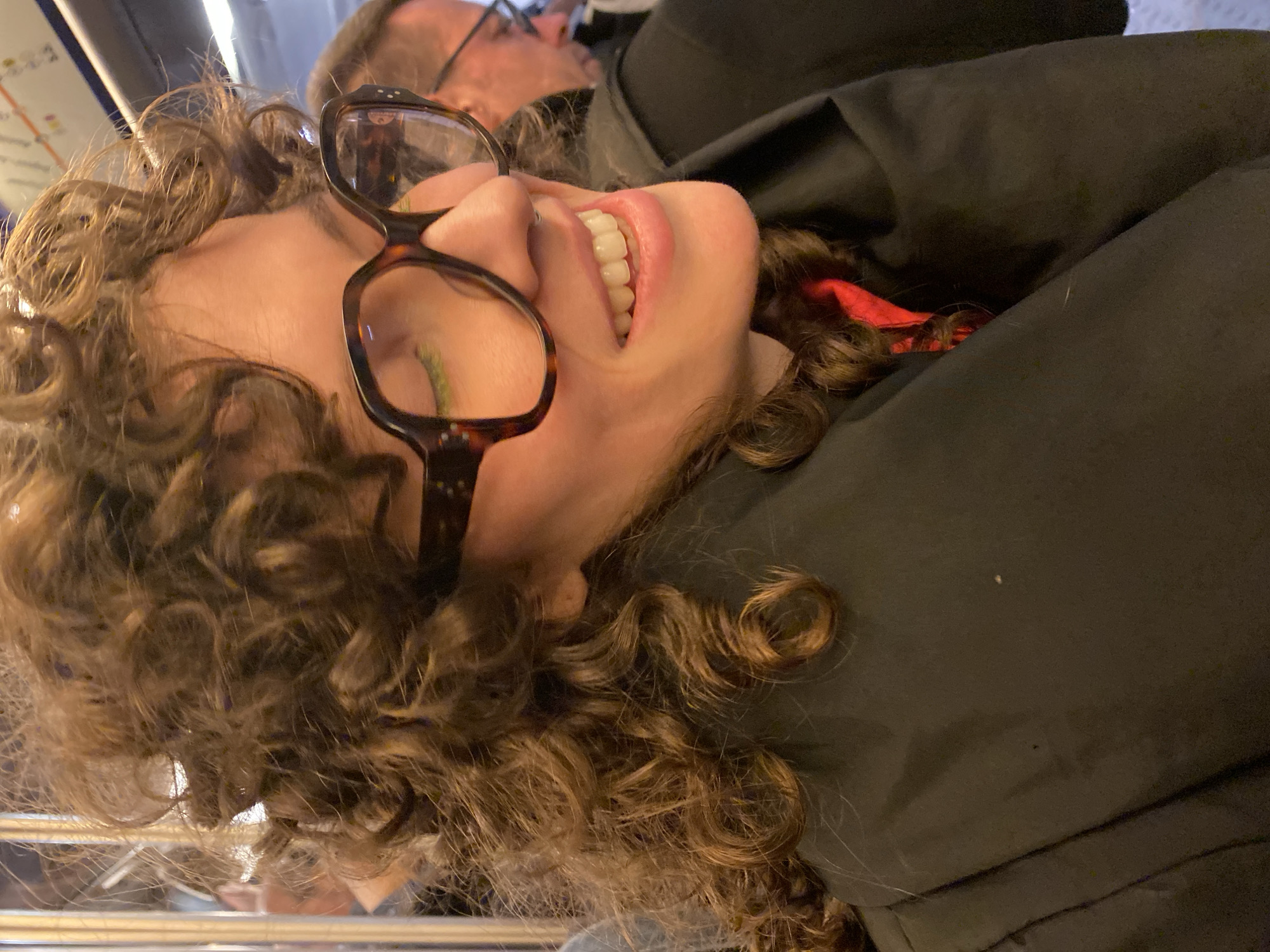 Close-up image of a smiling woman with curly hair and glasses. She is wearing a black jacket with a red scarf underneath. An older man with glasses is partially visible in the background. The location appears to be a public transportation vehicle, likely the Paris Métro, due to the presence of a map in the upper left corner.