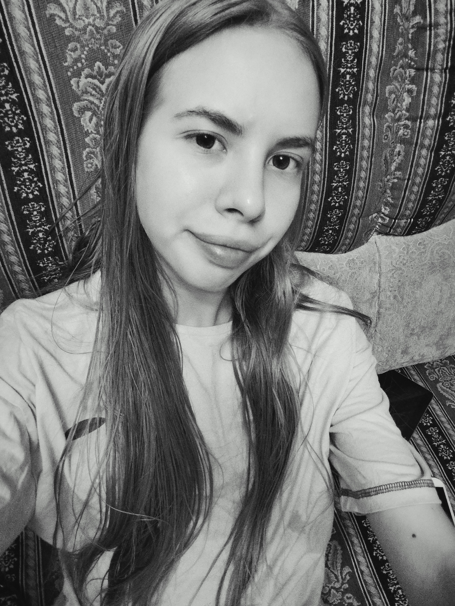 A black and white selfie of a young woman with long, straight hair is shown. She is looking directly at the camera with a slight smile. She is wearing a t-shirt. The background appears to be a patterned couch. The image is taken indoors.