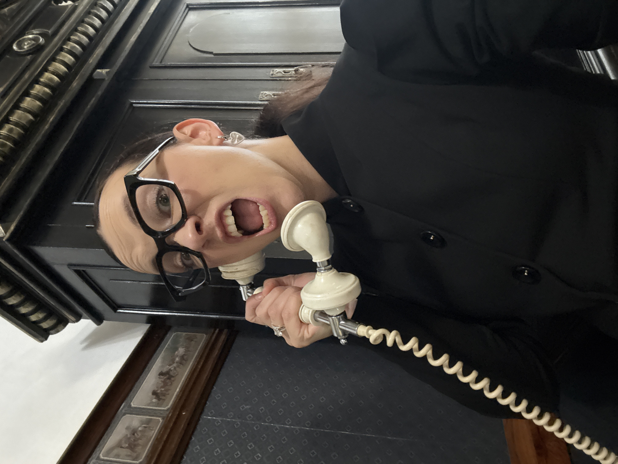 In Zavod Mikhailovskii, Russia, a woman with dark hair and glasses is holding an antique telephone to her ear and shouting into it. She is wearing a black jacket and silver heart-shaped earrings. The background consists of a dark wooden cabinet and a wall with a patterned wallpaper and framed artwork.
