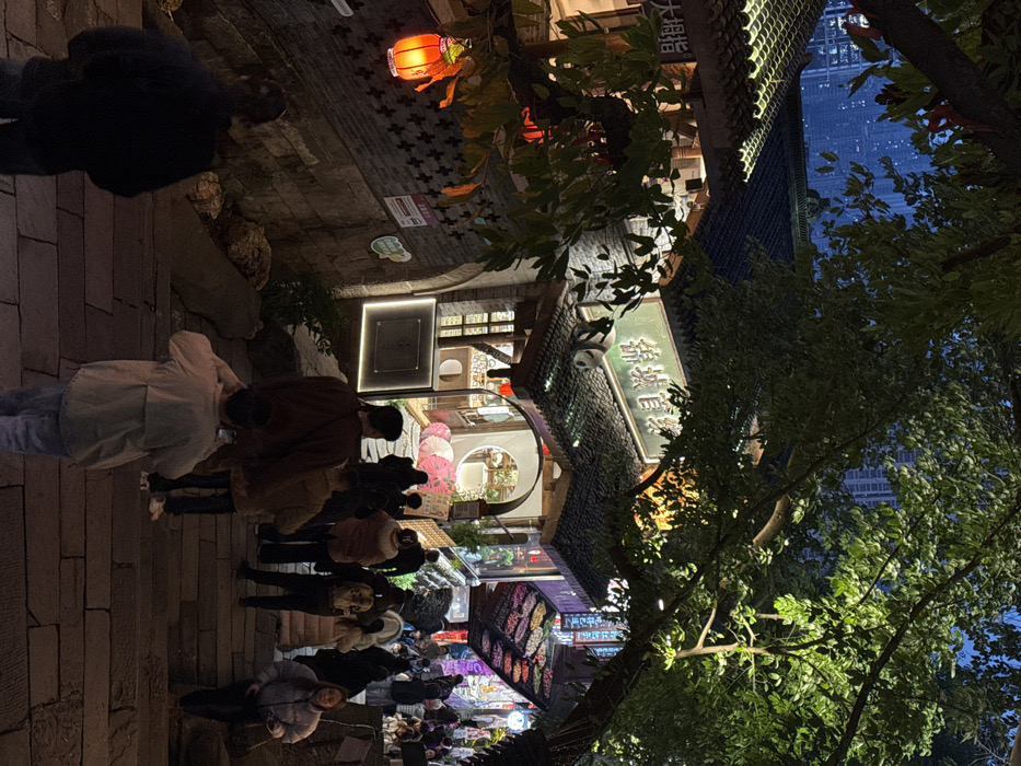 The image depicts a crowded outdoor staircase ascending a hillside at night in Yuzhong District, Chongqing, China. The scene is illuminated by artificial lights emanating from storefronts and lanterns.
The foreground features multiple individuals walking uphill on wide stone steps. A person in a light-colored jacket, with their back to the viewer, is holding a smartphone up, appearing to photograph or video the scene ahead. Other pedestrians, dressed in winter attire, are ascending the steps, some looking into storefronts.
To the left, a traditional-style building with a dark tiled roof and stone wall is visible. A prominent red lantern hangs on this wall. Above the building, a large green sign with Chinese characters reads "锦城官宴" (Jǐnchéng Guānyàn), and a black and white panda figure is situated on the roof beneath it. Another partially visible sign on the leftmost building reads "大拇指" (Dà Mǔzhǐ).
To the right of the main sign, another building features a circular arched entrance, revealing illuminated interior space with pink parasols or umbrellas arranged decoratively. Further right, a vibrant storefront displays rows of colorful merchandise under bright lights.
Large trees with green foliage are interspersed throughout the scene, some branches extending over the pathway and buildings. In the distant background, above the traditional structures, the silhouette of modern high-rise buildings indicates an urban environment. The sky is dark, consistent with nighttime.