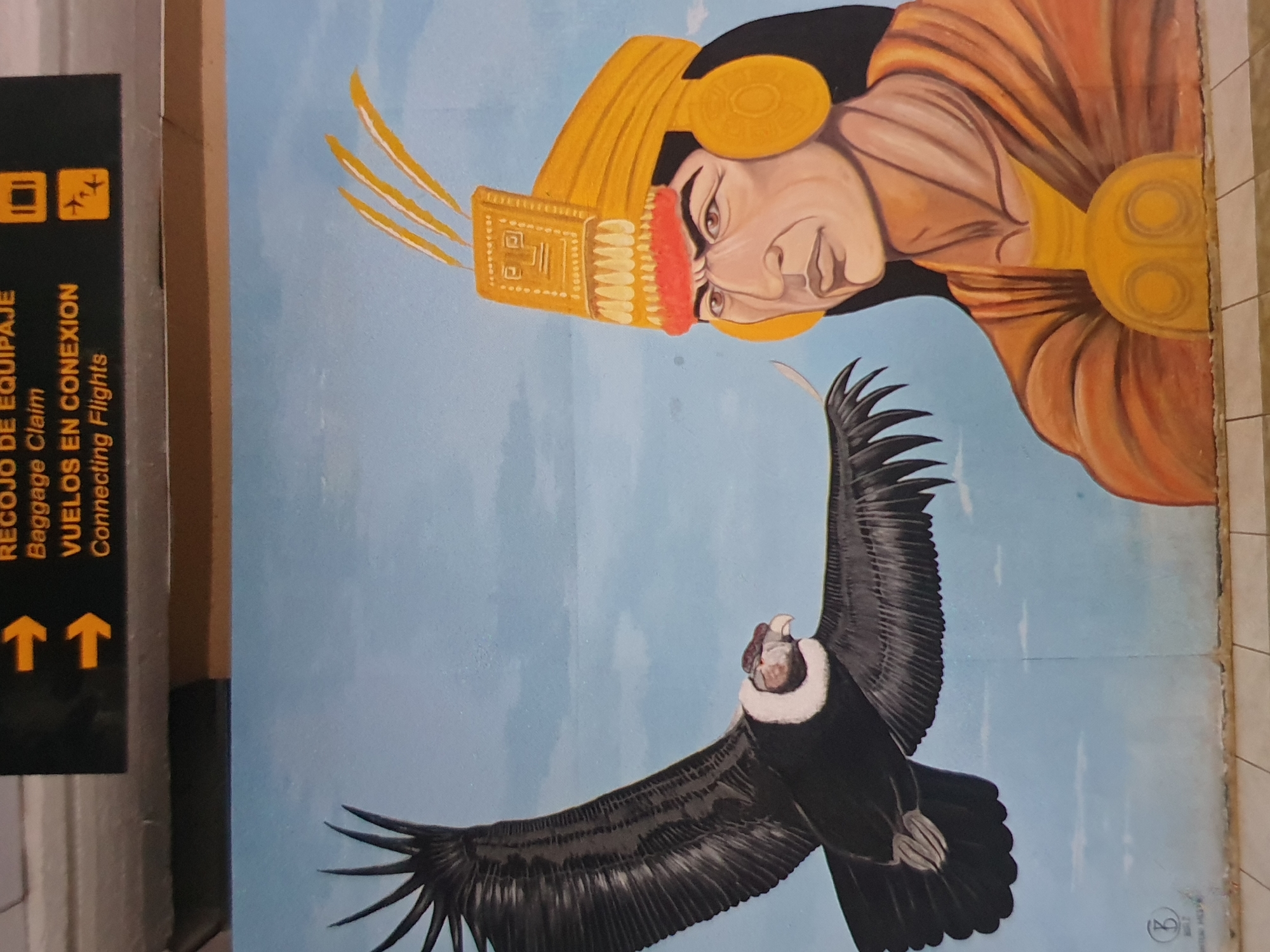 The image shows a mural in an airport, likely in South America. The mural depicts an Andean Condor flying below a portrait of an Incan ruler. The painting is stylized, with the condor having detailed feathers and the Incan figure wearing traditional headdress and attire. A sign with airport information in Spanish and English is adjacent to the mural, containing the words "RECOJO DE EQUIPAJE" (Baggage Claim) and "VUELOS EN CONEXION" (Connecting Flights), as well as directional arrows and airplane symbols. The mural is painted on a wall that appears to be slightly aged, with visible seams and wear.