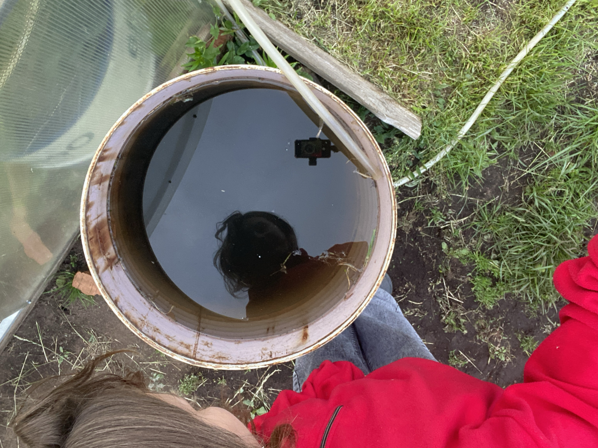 This image captures an outdoor scene in Chaikovsky, Russia, featuring a partial view of a person interacting with a large, partially filled barrel next to a greenhouse. The person, seen from an overhead perspective, wears a bright red long-sleeved top with a black zipper detail on the shoulder and light-colored trousers. Their brown hair is partially visible on the left. A large, rusty metal barrel, discolored with brown and reddish-brown stains, is positioned centrally. It contains dark, opaque water. The water's surface reflects the person's head and dark hair in the upper-left quadrant and a small, black camera (resembling a GoPro) mounted on a tripod in the lower-right quadrant. The camera's lens is visible, suggesting it is aimed upwards. To the upper right of the barrel, a section of a greenhouse constructed from translucent, corrugated panels is visible. The ground surrounding the barrel consists of dark soil, patches of green grass, and scattered dry plant debris. A piece of reddish-brown brick lies near the greenhouse. A translucent white plastic hose runs diagonally across the lower right portion of the image, extending towards and partially into the barrel's water. A weathered wooden plank rests on the ground parallel to the hose, adjacent to the barrel.