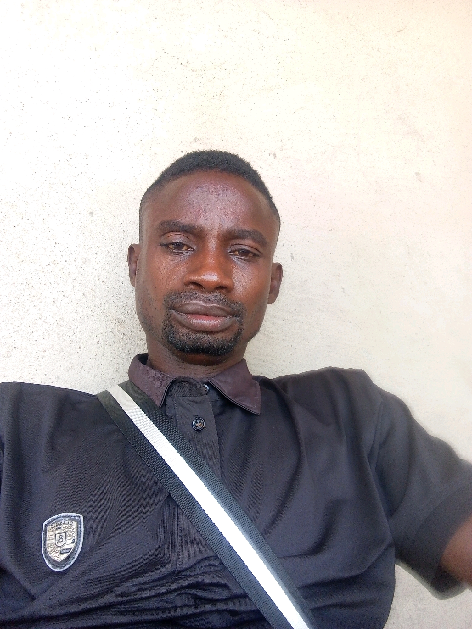 A medium close-up selfie shows a Black man in his late 20s or early 30s, with short dark hair and a beard, against a textured beige wall in Zango-ndaji, Nigeria. He is wearing a black polo shirt with a dark collar and a dark grey strap with white and black stripes crossing his chest. A shield-shaped logo with a number "8" is visible on the left side of his shirt. The man is looking directly at the camera with a neutral expression. The lighting suggests it is daytime.