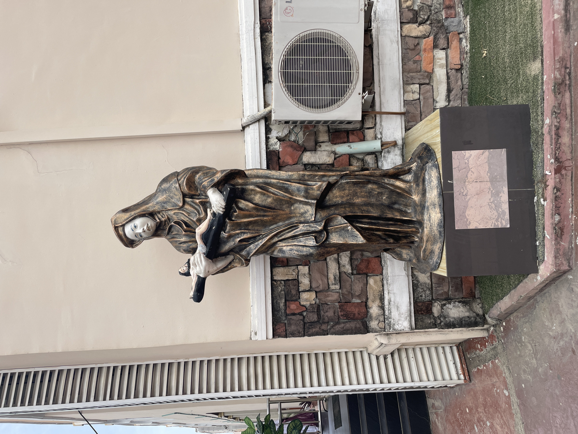 The image shows a statue of a cloaked figure holding a smaller figure, likely a religious sculpture depicting Mary holding Jesus. The statue is positioned on a pedestal beside a building with a cream-colored exterior wall, a brick section, and an air conditioning unit. In the foreground, there is a louvered structure and a section of the ground. The location is Unknown, Unknown.