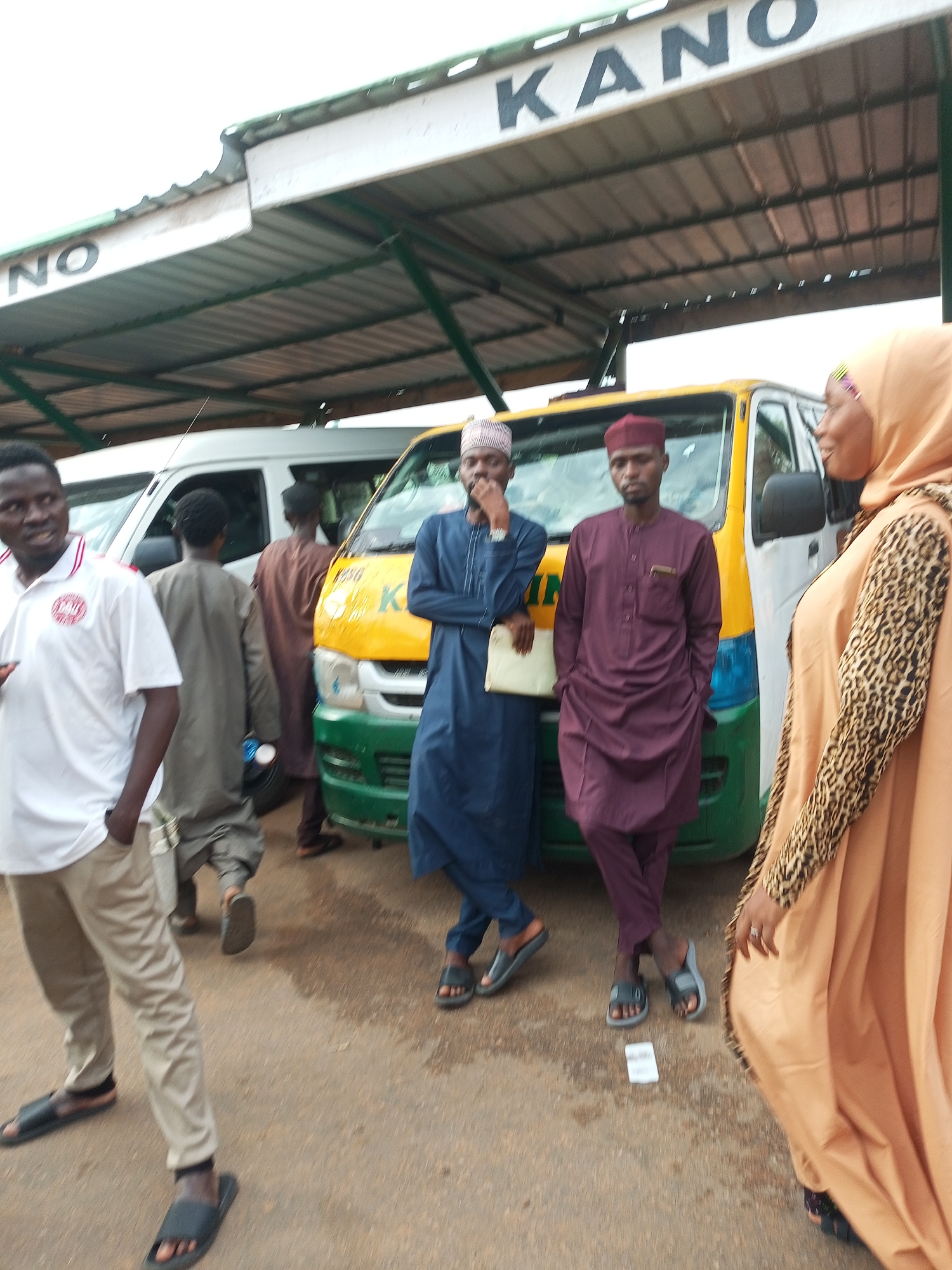 In Gombe, Nigeria, at a bus station, two men in traditional attire stand in front of a yellow and green bus. The man on the left wears a blue robe and cap, while the man on the right wears a maroon robe and cap. A woman in a light brown hijab and a leopard print top is partially visible to the right of the bus, facing away from the camera. Two other individuals, also in traditional clothing, are seen in the background, near another white bus. The scene is under a covered structure with a sign bearing the word "KANO" in large black letters. The ground appears to be asphalt and is wet, suggesting recent rain.