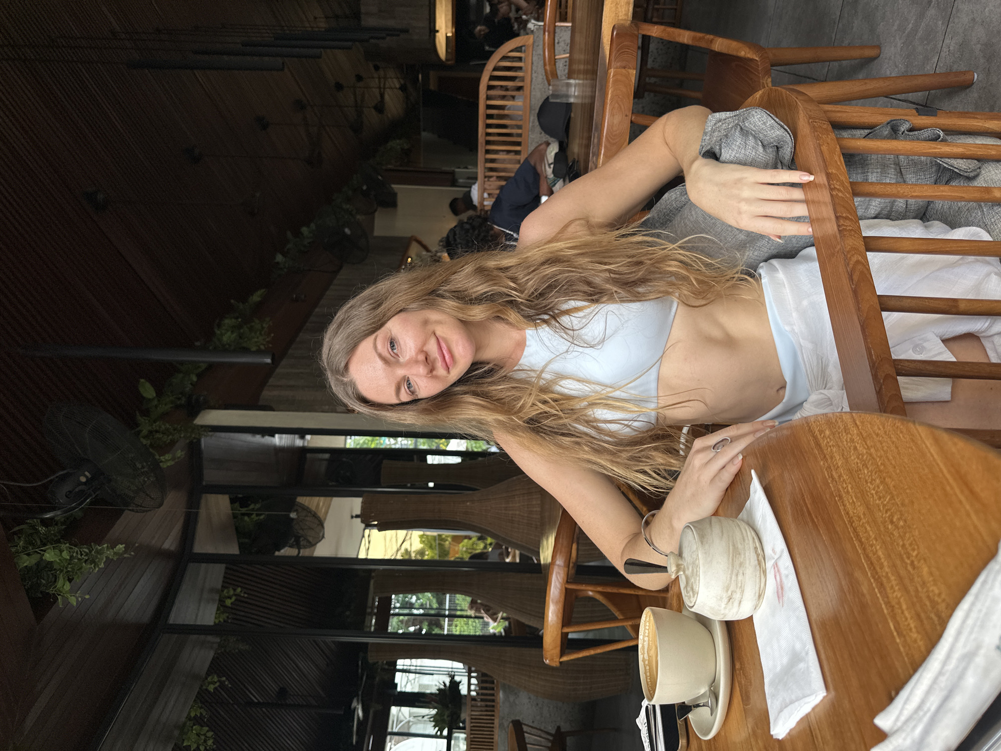 A woman with long, wavy blonde hair and a light blue top is seated at a wooden table in a cafe in North Kuta, Indonesia. She is looking at the camera and smiling. On the table in front of her is a cup of coffee, a sugar bowl, and a white napkin. Behind her, other patrons and decorative elements of the cafe are visible, including large woven vases and ceiling fans. The cafe appears to have a rustic, open-air design with wooden beams and seating.
