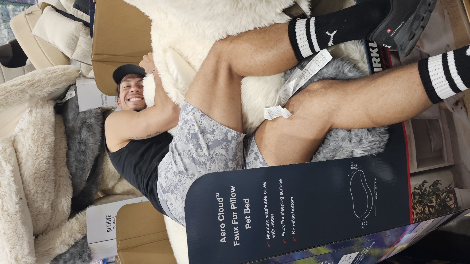 A young man with a wide, joyful smile is playfully posing amidst a large pile of faux fur pet beds and pillows, likely within a retail store or warehouse in Puebla, Mexico. He is captured reclining amongst the soft merchandise, with his body partially obscured by a large product box in the foreground. He is wearing a black tank top, light grey camouflage-patterned shorts, a black baseball cap, and black socks with white stripes. A Puma logo is visible on his left sock, and a black athletic shoe with "SWISS ENGINEERING" text is visible on his right foot.
The background and surrounding area are filled with numerous plush faux fur items in varying shades of cream, white, and grey, suggesting a display or stock of these products. Several cardboard boxes are visible, contributing to a cluttered but organized appearance of merchandise.
Prominently featured in the foreground is a large product box for an "Aero Cloud™ Faux Fur Pillow Pet Bed." Visible text on the box details its features: "Machine washable cover with zipper," "Faux Fur sleeping surface," and "Non-skid bottom." Dimensions are also listed as "81.2 cm / 32 in / 32 po" and "101.6 cm / 40 in / 40 po." Other discernible text on packaging includes "BEEHIVE 2-IN-1" and "KIRKL" (likely Kirkland). The man's happy expression suggests he is enjoying the comfort of the faux fur products. The scene is brightly lit, indicative of an indoor commercial setting.