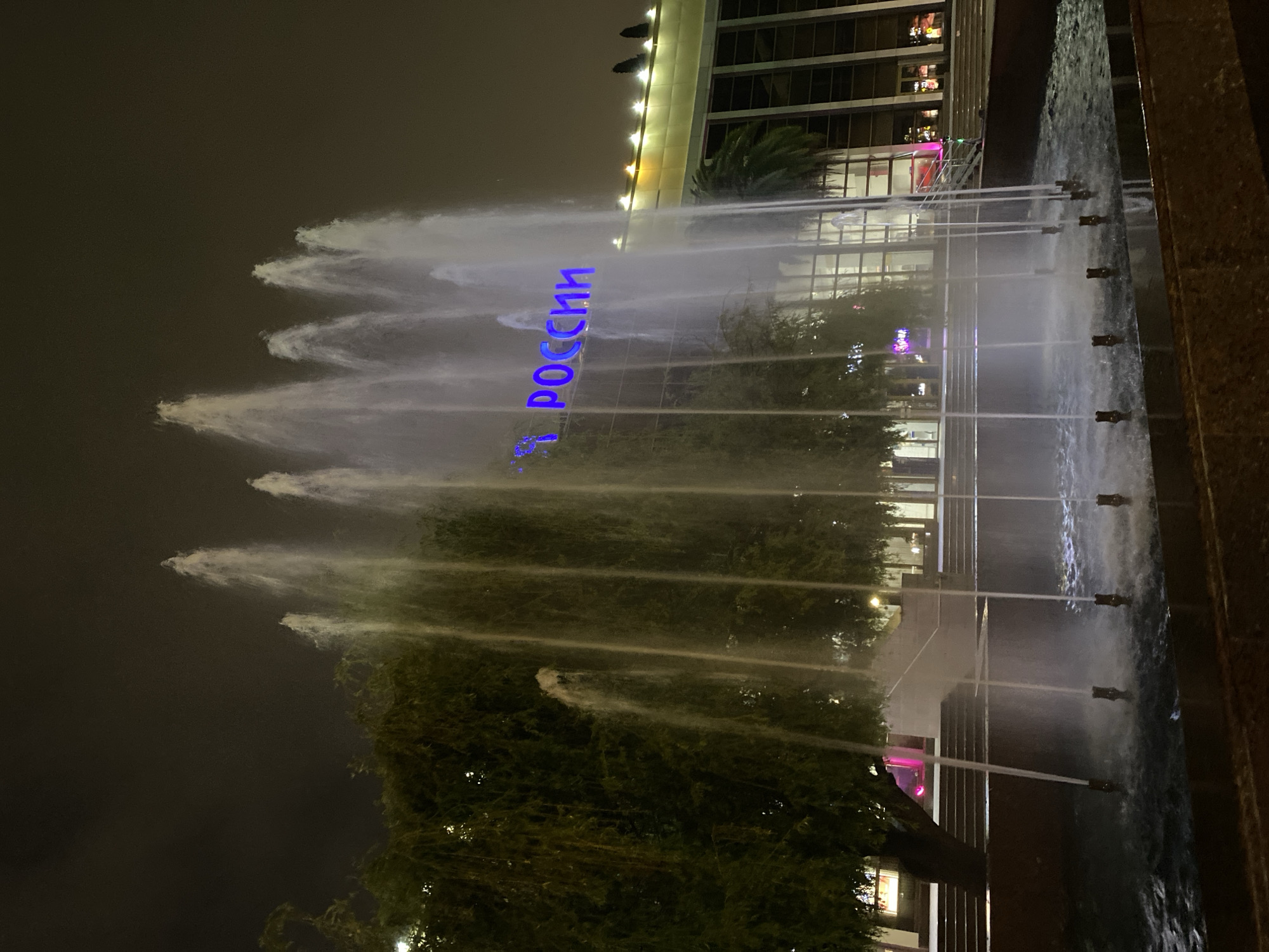 The image shows a nighttime view of a fountain in Sochi, Russia. The fountain jets water upwards in multiple streams, with the name "Россия" (Russia) illuminated in blue neon lights above the fountain, superimposed on the background building. The building is multi-storied with a modern architectural design, featuring numerous windows. A few trees are visible in the foreground, and the night sky is dark. The fountain is situated beside a walkway.