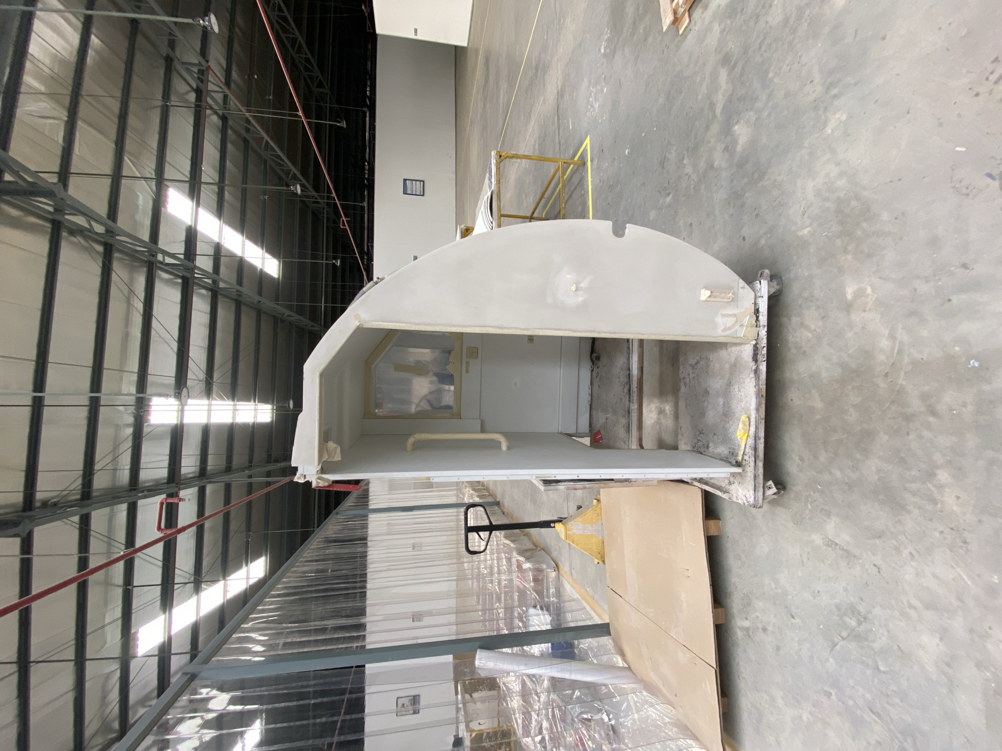 The image depicts the interior of a large industrial warehouse or factory floor in Batam, Indonesia. The space is open with a high ceiling supported by metal trusses. Natural light streams in from skylights in the roof, and artificial lights are also visible. The floor is concrete, showing signs of wear and dust.
The central focus is a partially assembled, white, modular structure, possibly a booth or a cabin, mounted on a wheeled platform. The structure has an opening with steps leading into it, and a small window is visible. To the left of the structure, a yellow pallet jack is positioned near some large cardboard rolls wrapped in plastic. Further to the left, transparent plastic sheeting hangs, dividing the space.
In the background, the warehouse extends with rows of stacked items, also covered in plastic. A yellow metal table with some papers on it is situated in the middle ground. The overall scene suggests an ongoing manufacturing or assembly process within an industrial setting. There are no people visible in the image.