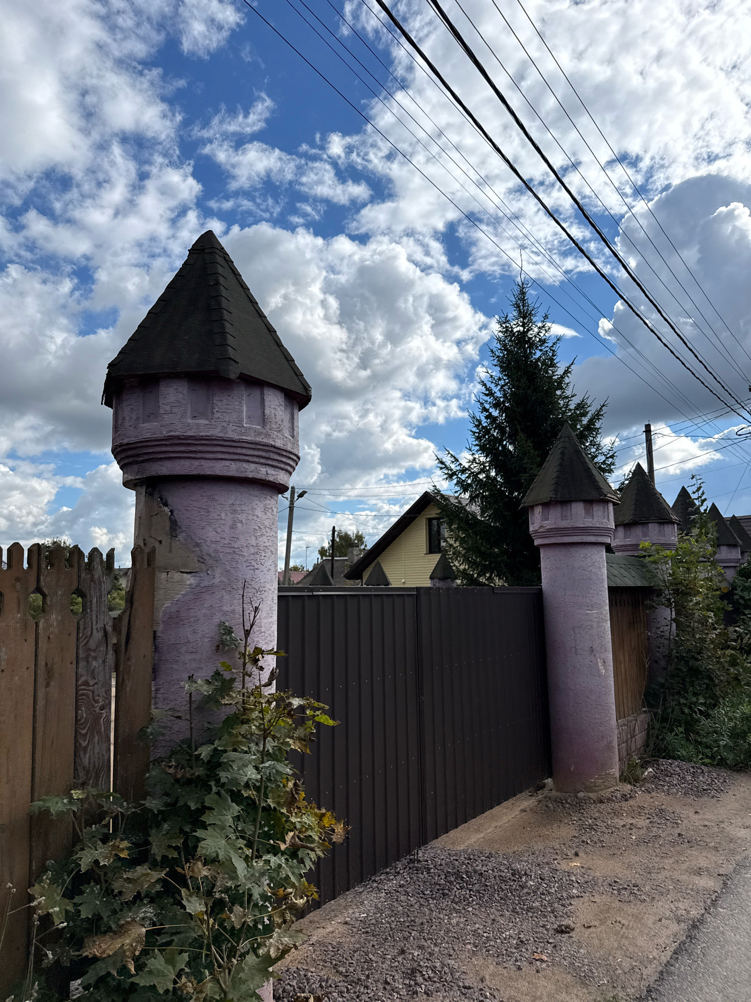 The image shows a residential street in Vsevolozhsk, Russia, with a cloudy blue sky overhead. The foreground features a wooden fence and a metal gate, flanked by two turret-like structures painted lavender. These structures have dark, conical roofs and decorative battlements. Behind the gate, a tall evergreen tree stands out against the sky, with houses and more turrets visible in the distance. Power lines run diagonally across the upper portion of the frame. The ground is covered with gravel and dirt.