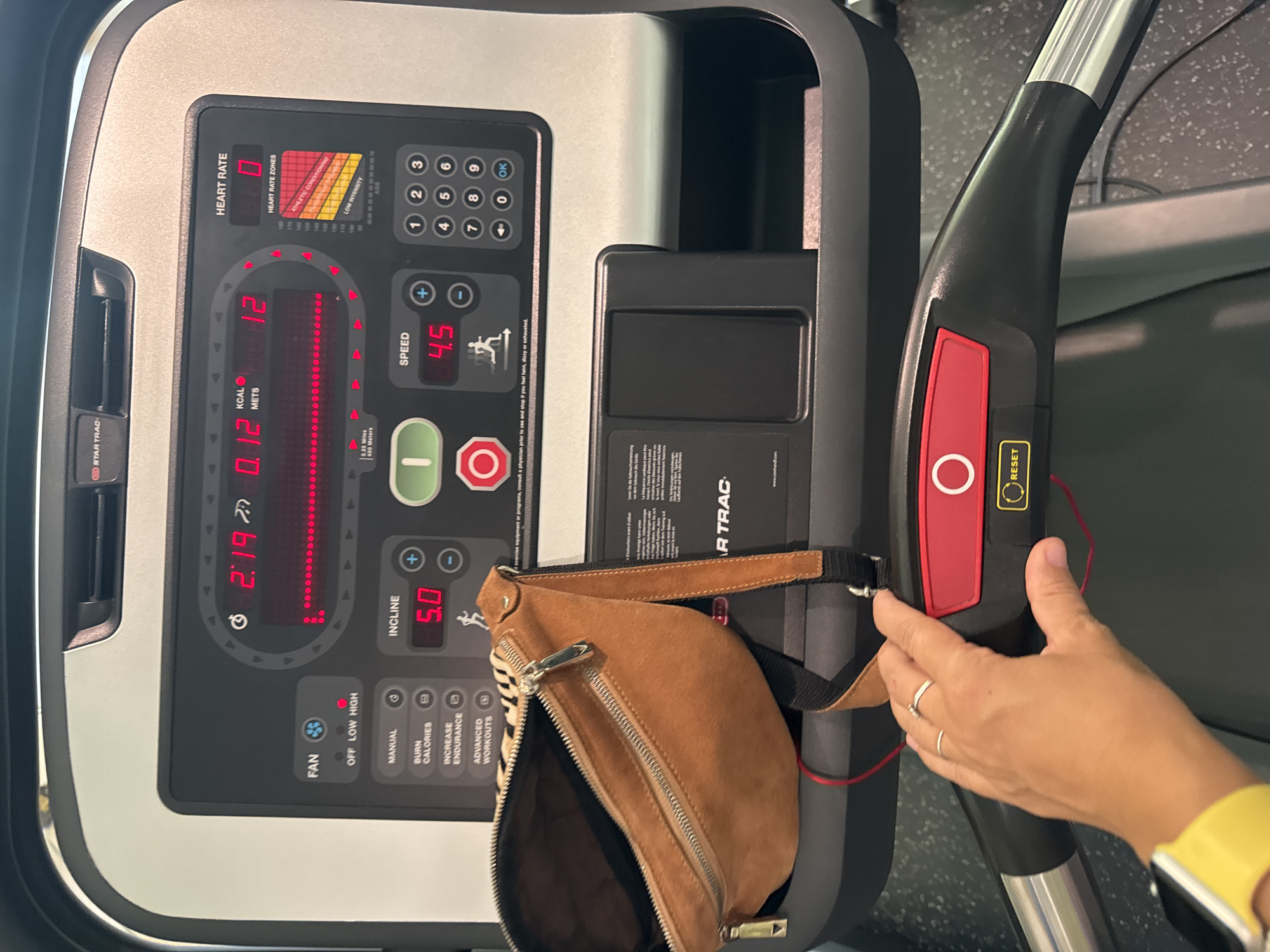 The image shows a close-up view of a treadmill console in Kuta Selatan, Indonesia. The console displays various metrics including time, calories, heart rate, incline, and speed. The time is 2:19, incline is set to 5.0, and speed is 4.5. There is also a heart rate reading of 0 and a heart rate zone indicator. A tan fanny pack is placed on the treadmill's deck. A person's hand, adorned with two rings, is visible in the foreground, touching the treadmill's handlebar which has a red "RESET" button. The environment appears to be a gym, with a speckled floor visible.