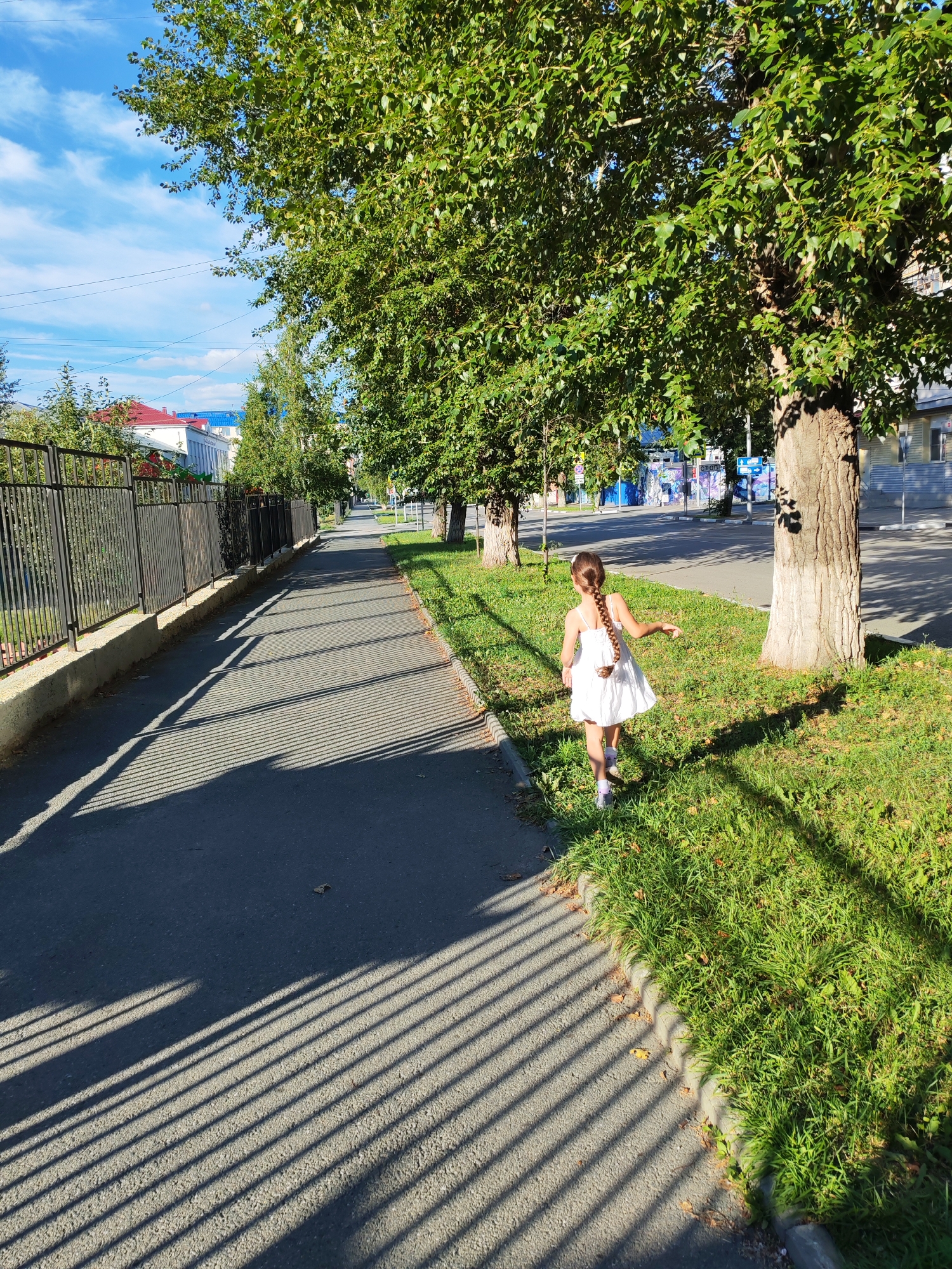 A young girl with a braided pigtail, wearing a white dress and pink shoes, walks on the grass beside a sidewalk lined with trees. She is facing away from the camera and looking to her right. The sidewalk is shaded by the trees, casting long, striped shadows. A black metal fence runs along the left side of the sidewalk. In the background, buildings and a street with a few cars are visible. The sky is blue with scattered white clouds.