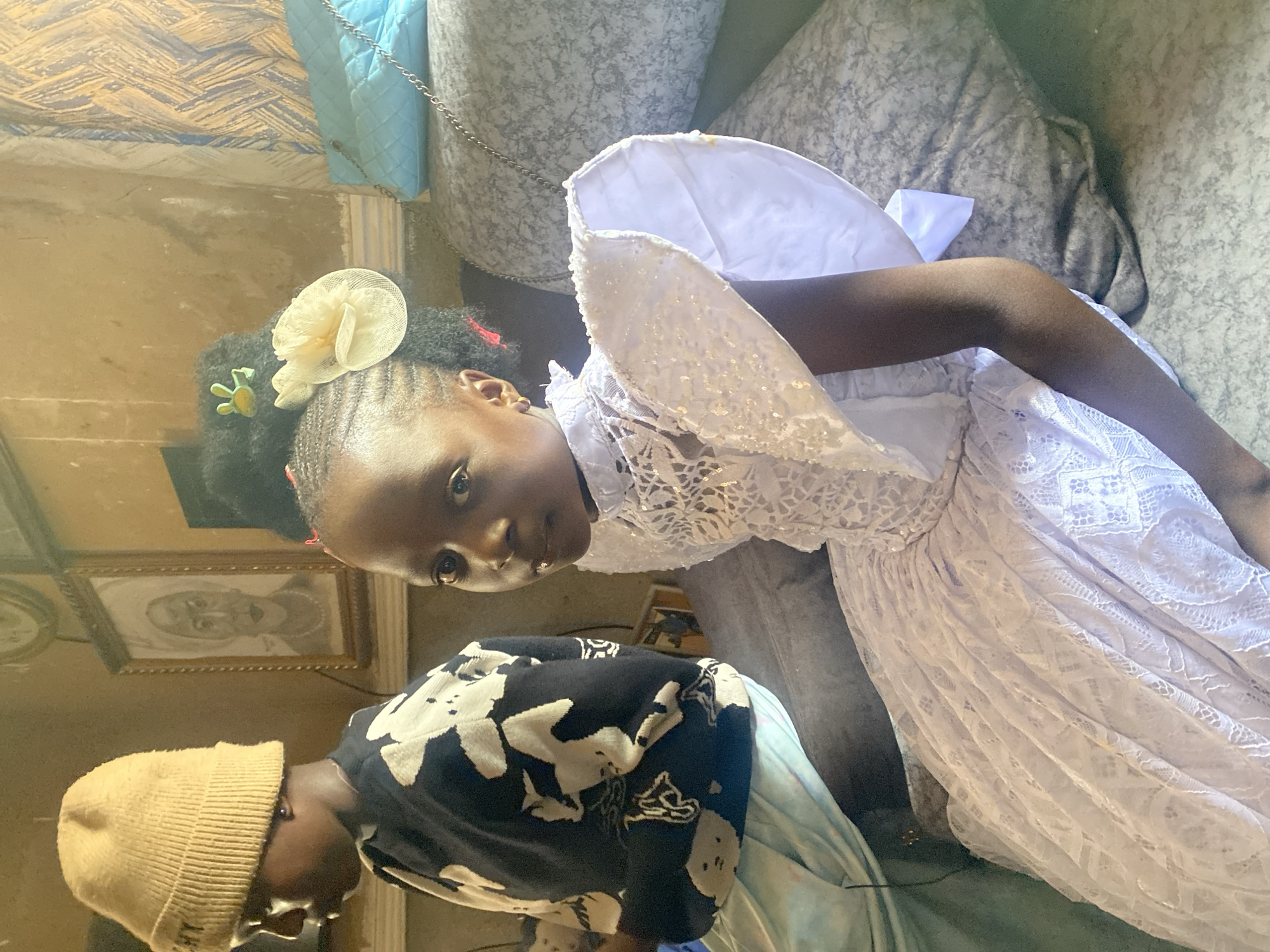 A young girl in a white lace dress and angel wings lies on a gray patterned couch. She has a white and yellow flower in her hair. Another child wearing a tan beanie and a black and white patterned shirt stands next to her. On the wall behind the child, there is a framed portrait. The location is Kuru Dungus, Nigeria.