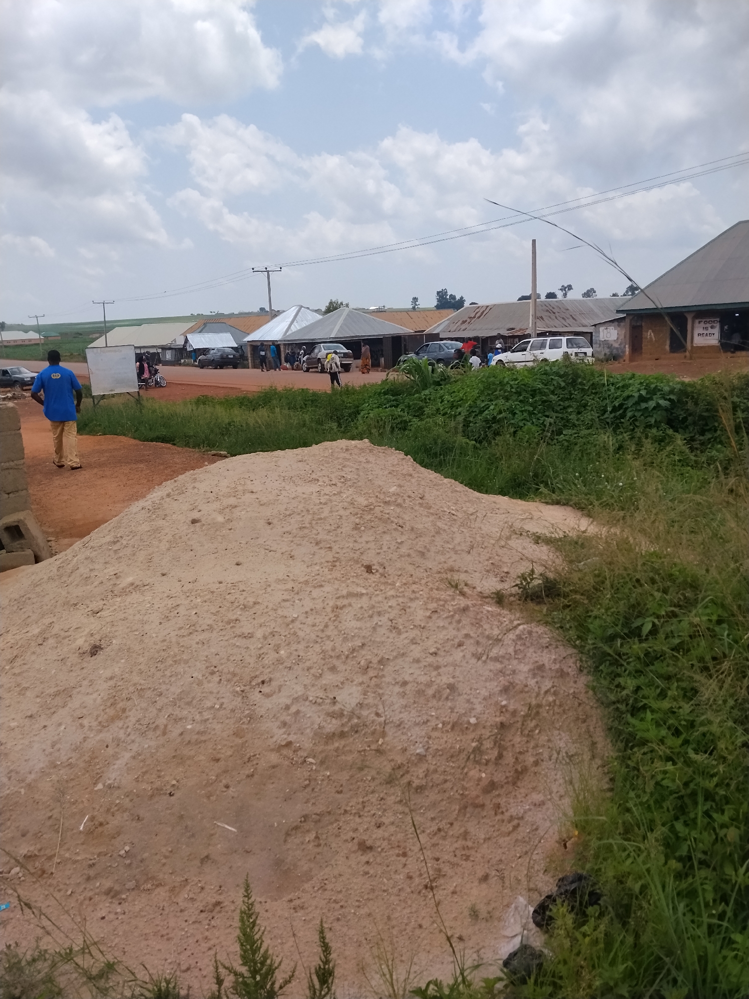 The image depicts a roadside scene in Durumi, Nigeria. In the foreground, a large pile of light brown sand dominates the lower half of the frame. To the left and slightly behind the sand pile, a man wearing a blue shirt and tan pants walks away from the camera.
Further back, a dirt road stretches into the distance, lined with various market stalls and buildings with corrugated metal roofs. Several vehicles are parked along the road, including a white SUV and a dark sedan. A few people are gathered around the stalls, some appearing to be vendors and customers.
The environment is characterized by a cloudy sky and sparse vegetation growing along the sides of the road and behind the market structures. A white sign with text is visible attached to one of the market stalls. The overall impression is of a bustling, informal marketplace in a rural or semi-urban setting.