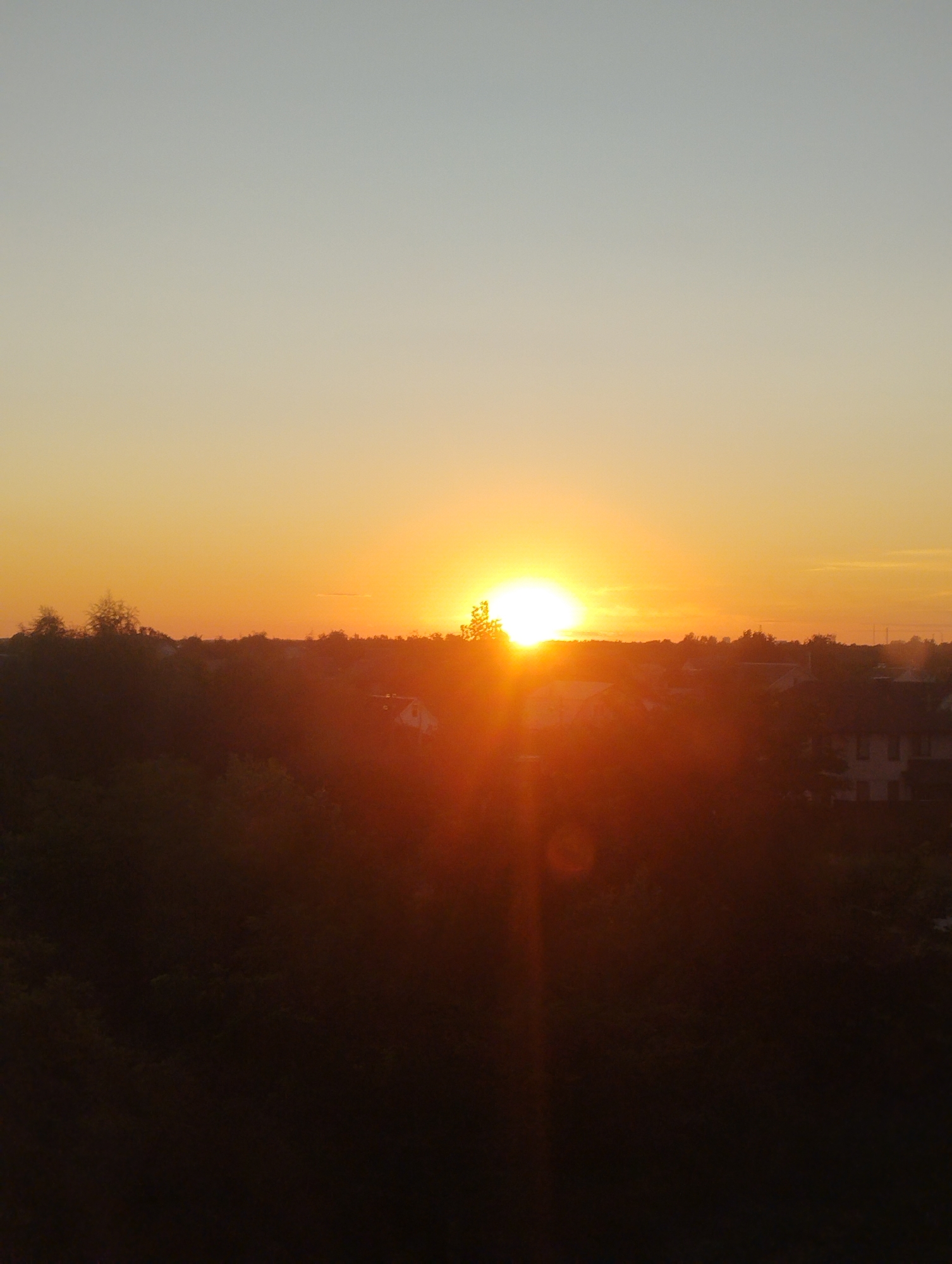A vertical, medium-shot photograph captures a sunset over a residential area in Homyel, Belarus. The sky transitions from a pale blue at the top to a vibrant orange near the horizon, where the sun is partially obscured by trees and buildings. Silhouetted against the bright sky are the dark shapes of trees, rooftops, and the faint outline of houses. A strong lens flare emanates from the sun, creating a bright, hazy glow that obscures many of the finer details of the landscape. The overall impression is one of a peaceful, early evening scene.