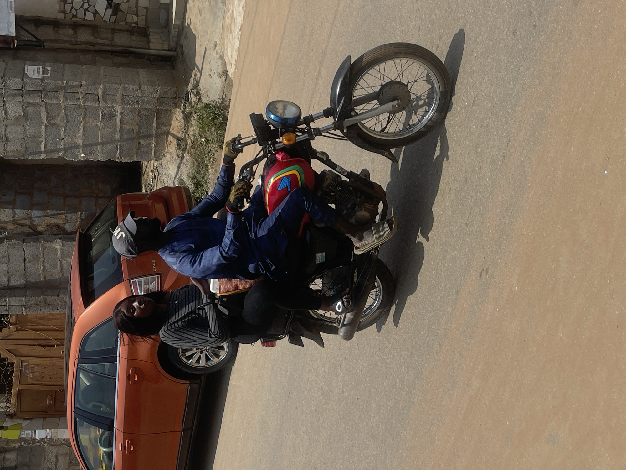 An African man wearing a blue tunic and dark cap, and an African woman wearing a striped long-sleeved shirt and black leggings, are riding on a red and yellow Honda motorcycle on a street in Abuja, Nigeria. The man is driving the motorcycle, holding the handlebars and wearing gloves. The woman is a passenger, sitting behind the man, holding onto him. A red car is parked on the left side of the street, behind the motorcycle. The background shows a brick wall and some vegetation. The street appears to be paved with asphalt.