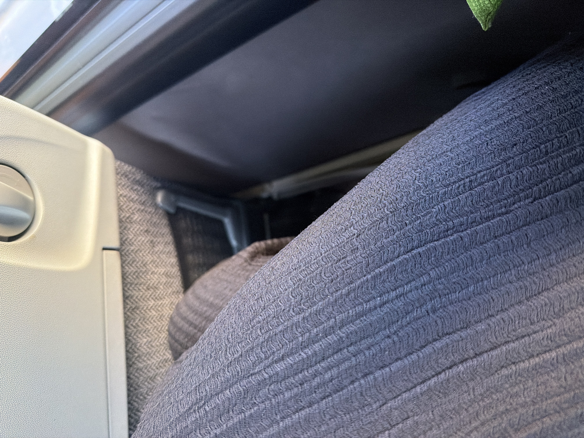 The image shows a close-up view of a person's legs covered in blue ribbed fabric, likely trousers or a skirt, sitting in what appears to be a vehicle's interior. The fabric has a distinct zig-zag pattern within the ribbing. To the right of the legs, a dark, textured surface is visible, possibly the interior of a car seat or panel. A black, lever-like object is partially visible between the person's legs and the dark surface. Above the fabric, a light-colored, textured surface, possibly a car's interior panel or armrest, is seen with a circular, metallic-looking object embedded in it, suggesting a control or vent. A portion of a window or reflective surface is visible to the far right, showing a bright, out-of-focus background. The scene is indoors, likely within a public transport vehicle or private car, but no specific location in Plovdiv, Bulgaria, can be determined from the visual information provided.