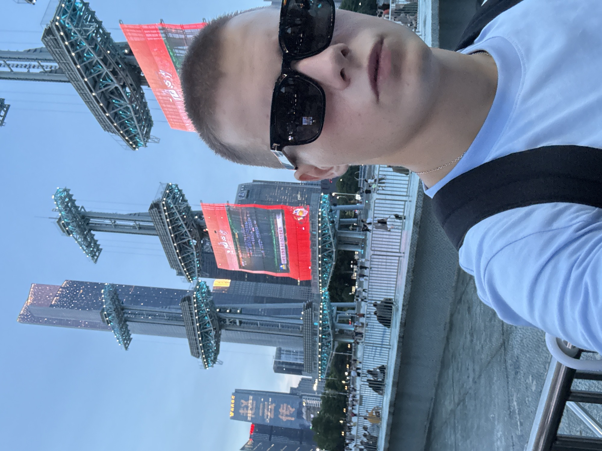 This media file captures a selfie taken by a young person wearing sunglasses and a light blue t-shirt, with a black backpack strap visible. The person is looking towards the camera. The scene appears to be outdoors in a city at dusk, with the sky showing a clear, pale blue hue, suggesting good weather.
In the background, a modern urban landscape is visible. Tall buildings, some illuminated with sparkling lights, dominate the skyline. Large, illuminated structures resembling stages or event setups with scaffolding and lights are prominent, one featuring a large red banner with Chinese text. There are also red banners with Chinese characters visible on some buildings. The presence of these structures and banners, along with a crowd of people gathered on a bridge or elevated walkway in the middle ground, suggests an outdoor event or performance is taking place. The mood seems festive and vibrant, indicated by the illumination and the gathering of people. The location is identified as Tianhe District, China, which aligns with the urban environment and the visible Chinese text.