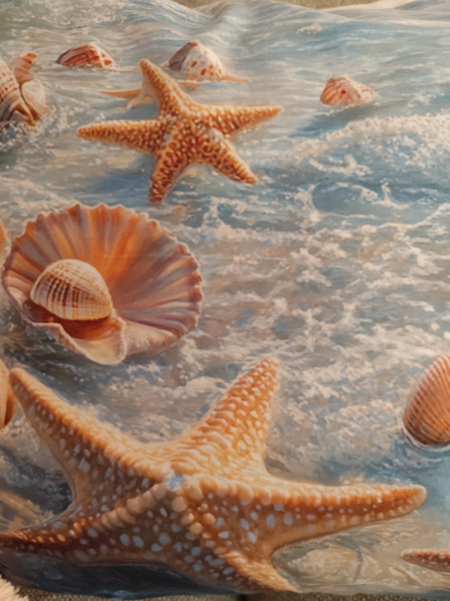 The image depicts a close-up view of a fabric print featuring seashells and starfish amidst gentle waves. Several seashells, in shades of brown and white, are partially submerged in the blue water. Two prominent starfish, one in the foreground and another in the mid-ground, are rendered in warm orange hues with white speckled detailing. A large, ribbed seashell, likely a scallop, is centrally positioned, cradling a smaller, striped shell. The water is characterized by white foam and ripples, suggesting a shallow, calm sea. The overall impression is one of a serene beach scene. The location is not a city in Montana, Bulgaria, as the image depicts a fabric print and not a real-world scene.