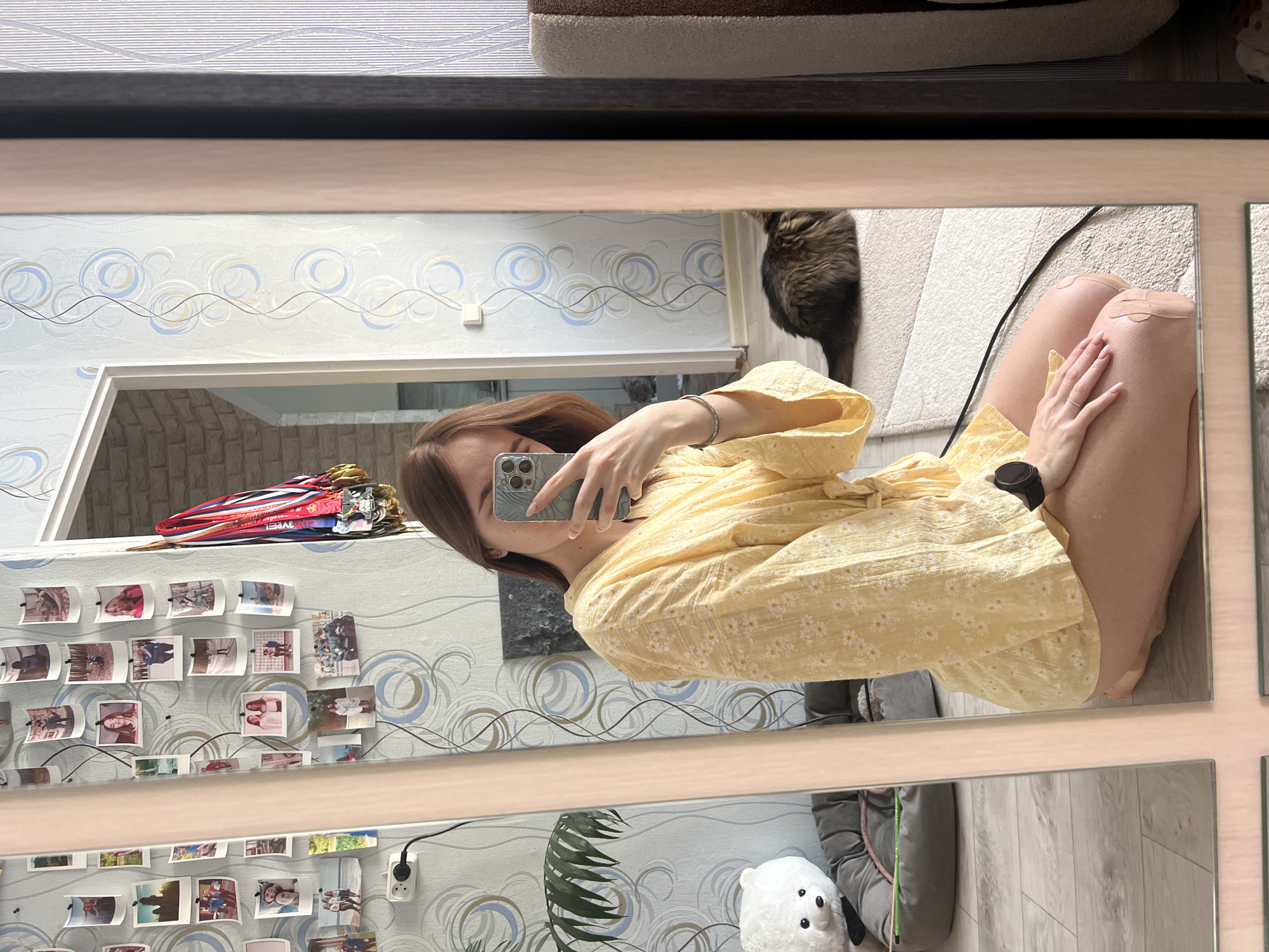 A forensic description of the scene within a residence in Chaikovsky, Russia, shows a female subject taking a mirror selfie.
**Subject:** A light-skinned female, partially obscured, is kneeling on a light beige shaggy rug. She is wearing a yellow, short-sleeved robe patterned with small white daisies, tied at the waist. Her left hand, with manicured nails and a silver ring on the ring finger, holds a light-colored smartphone (likely an iPhone) to capture the image, obscuring her face. A black smartwatch is visible on her right wrist. Two light-colored adhesive bandages are applied to her right knee. Long, light brown hair is visible on her left side.
**Environment (Reflected in Mirror):** The room features light blue/grey wallpaper with a repeating pattern of white, blue, and gold concentric circles and wavy lines. Numerous small, printed photographs, primarily portrait-oriented and depicting individuals or groups, are attached to the wallpaper on the left. A white electrical outlet with a black cord is visible on this wall. To the left of the subject, a white plush teddy bear sits in a grey pet bed containing a green toy stick with a pink tip, all positioned on a light wood laminate floor. Further back, a green leafy houseplant is visible. To the right of the subject, a fluffy brown and black cat is lying on the beige shaggy rug. Adjacent to the wallpaper on the right, a collection of sports medals with red, white, and blue ribbons is hanging. A white door frame opens to an adjacent room with light-colored, brick-patterned wallpaper. On the far right, a section of striped brown and beige upholstered furniture is partially visible. The lighting is bright and even, suggesting daytime.