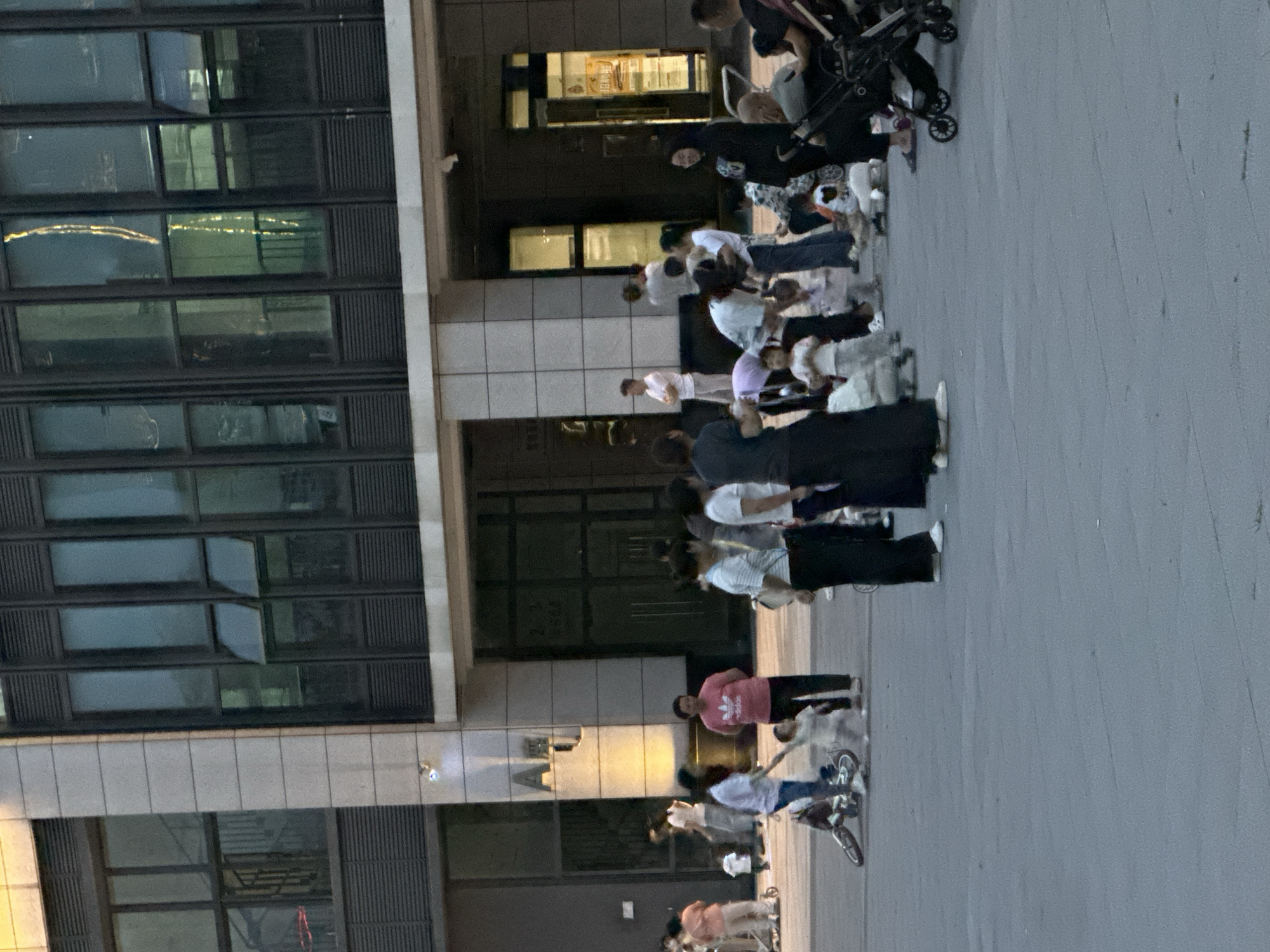 The image depicts an outdoor scene in the Chongqing High-tech Zone, China, likely a public square or pedestrian area adjacent to a modern building. Several individuals, including adults and children, are present. Some adults are standing, talking, or supervising children. Children are engaged in various activities, such as playing with a bicycle, crawling on the ground, or being held by adults. Some individuals are pushing strollers. The building in the background features large windows and a contemporary architectural style. The ground is paved, and the lighting suggests it is late afternoon or early evening.