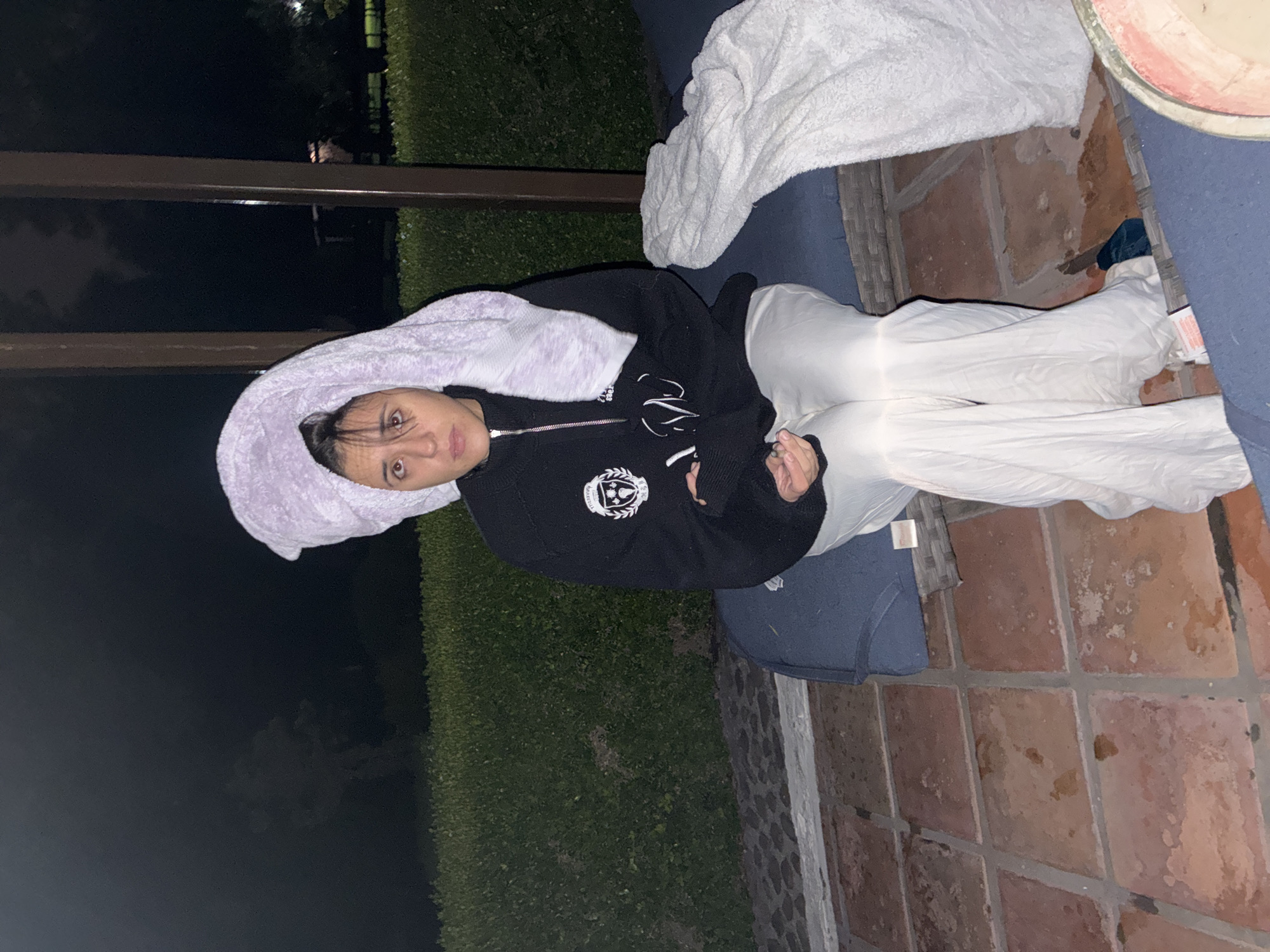 A woman with a lavender towel on her head is sitting on a blue outdoor couch on a patio. She is wearing a black jacket and white pants. The patio is made of red bricks. There is a bush in the background. The location is presumably Cañada de Madero, Mexico. It is nighttime.