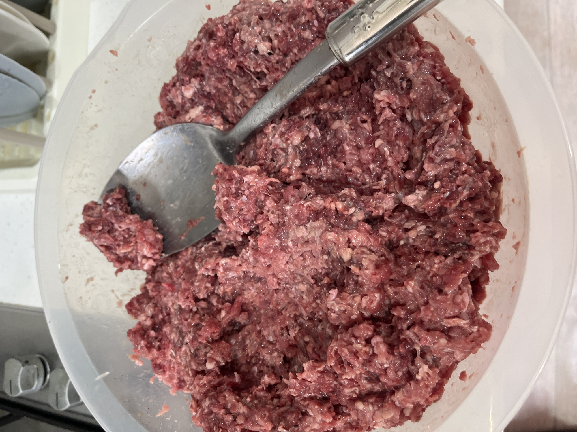 A white plastic bowl is filled with a large quantity of ground meat. A metal spoon is resting within the bowl on top of the meat. The bowl is on a counter top, and the corner of a stovetop appliance is visible in the lower left. The location is unknown and cannot be ascertained from the image.