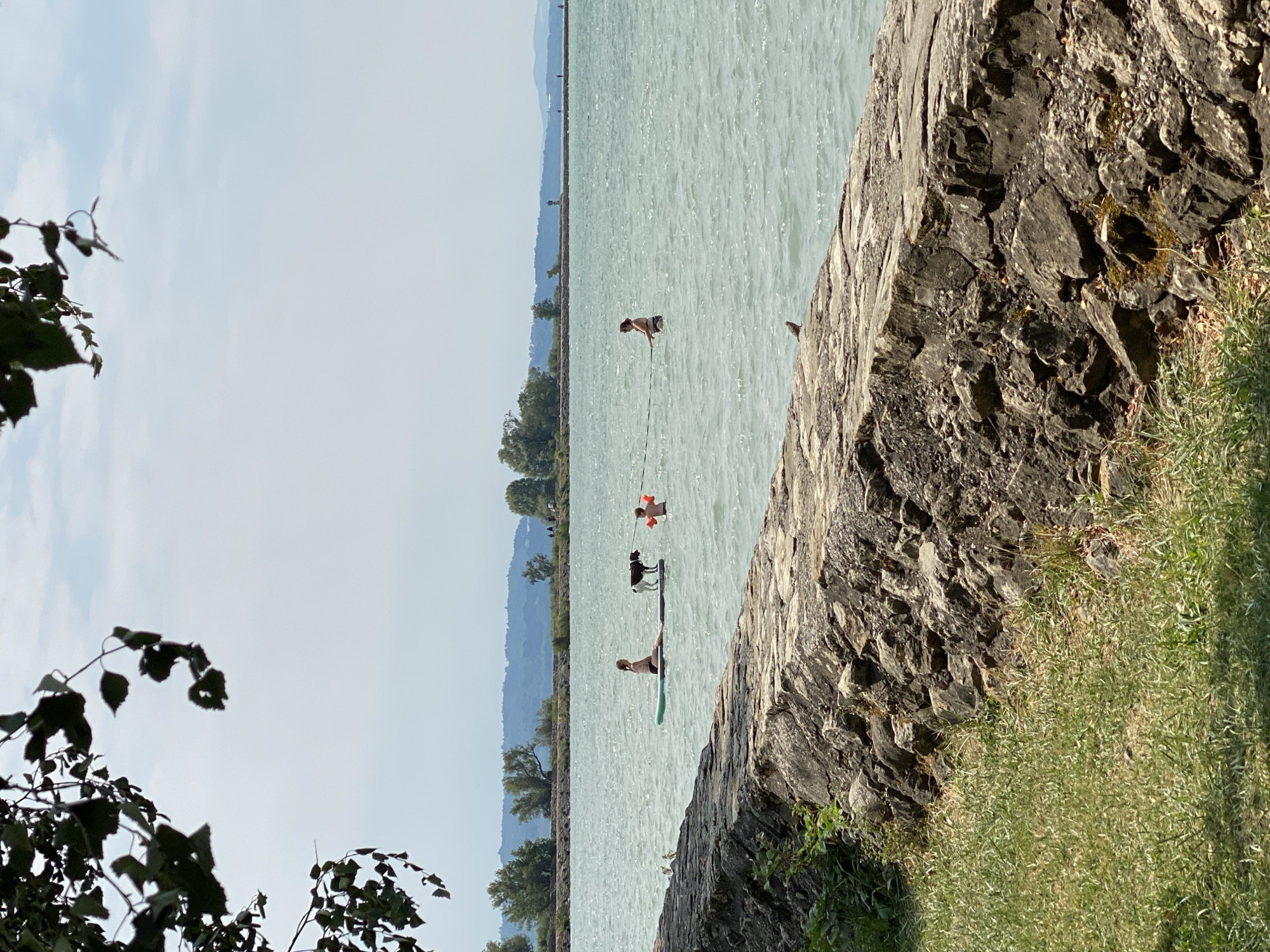 The image depicts a sunny day at a body of water, likely a lake, in Laténa, Switzerland. In the foreground, a stone embankment lines the grassy shore. In the middle ground, a pale blue lake stretches out, with several people and a dog present. A woman is paddleboarding with a dog on the board. Nearby, a child in an orange flotation device is in the water. Further out, another woman is standing in the water, holding onto a rope. In the background, trees and hills are visible, with a hazy sky overhead.