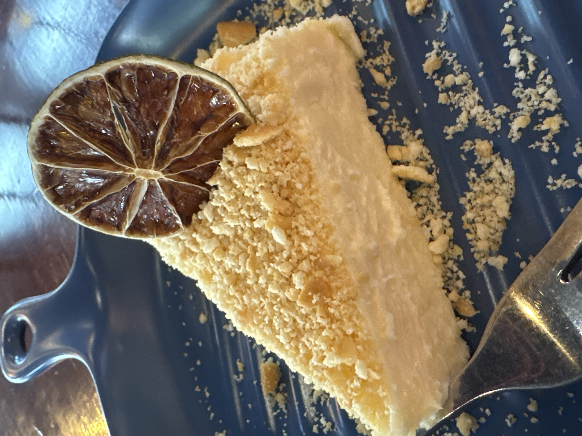 A close-up photograph shows a slice of cake on a blue plate. The cake is rectangular in shape and has a creamy, white frosting. It is covered in crumbled biscuit crumbs. A dried lime slice adorns the cake. A fork is visible on the right side of the plate. The plate is blue and has a handle on the left side. Fine crumbs are scattered on the plate. The background appears metallic or reflective. Location information is not discernible.